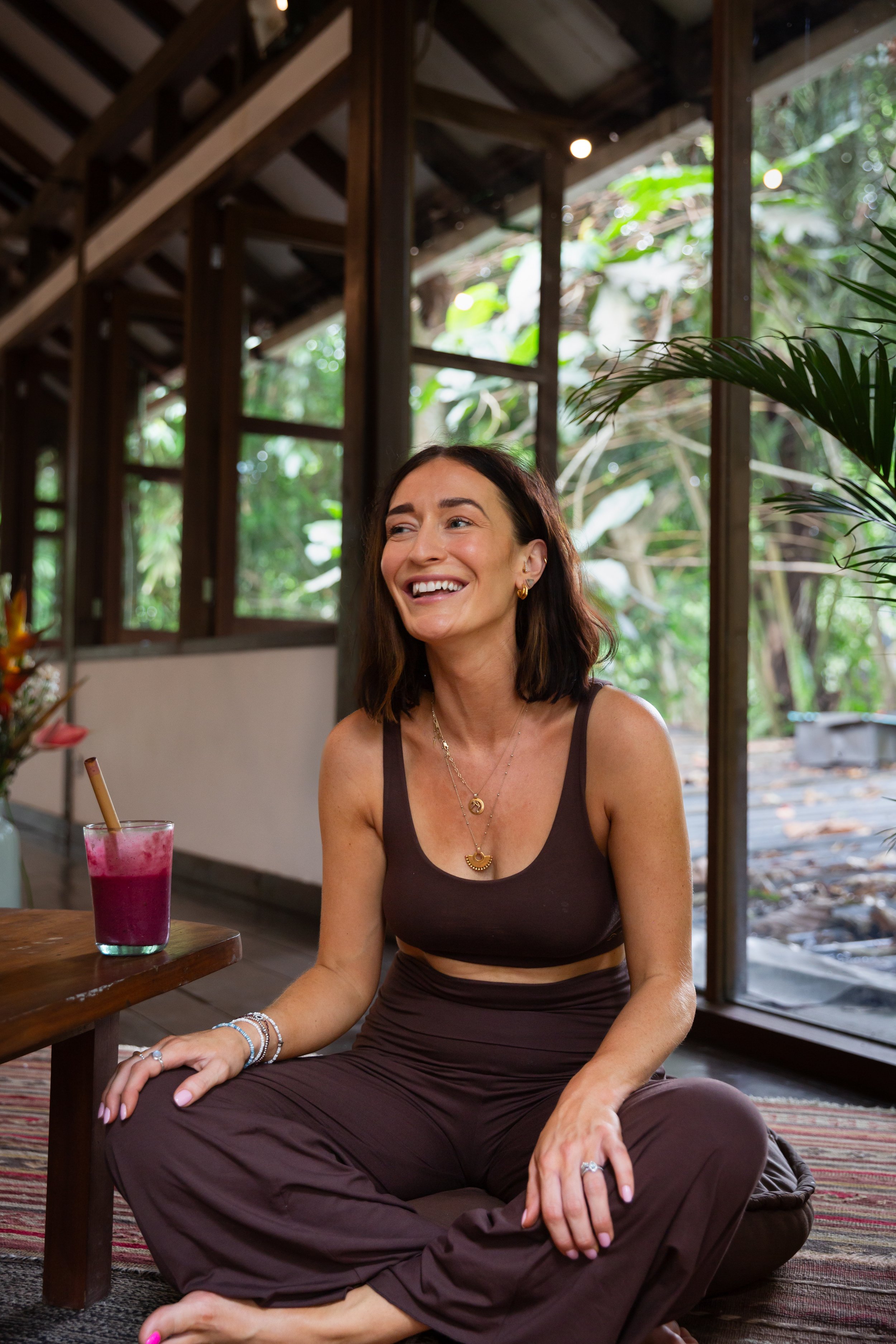 A woman sitting on the floor in a relaxed indoor setting, wearing a brown outfit and smiling. There is a glass of pink smoothie on a wooden table beside her. The background shows large windows with a view of greenery.
