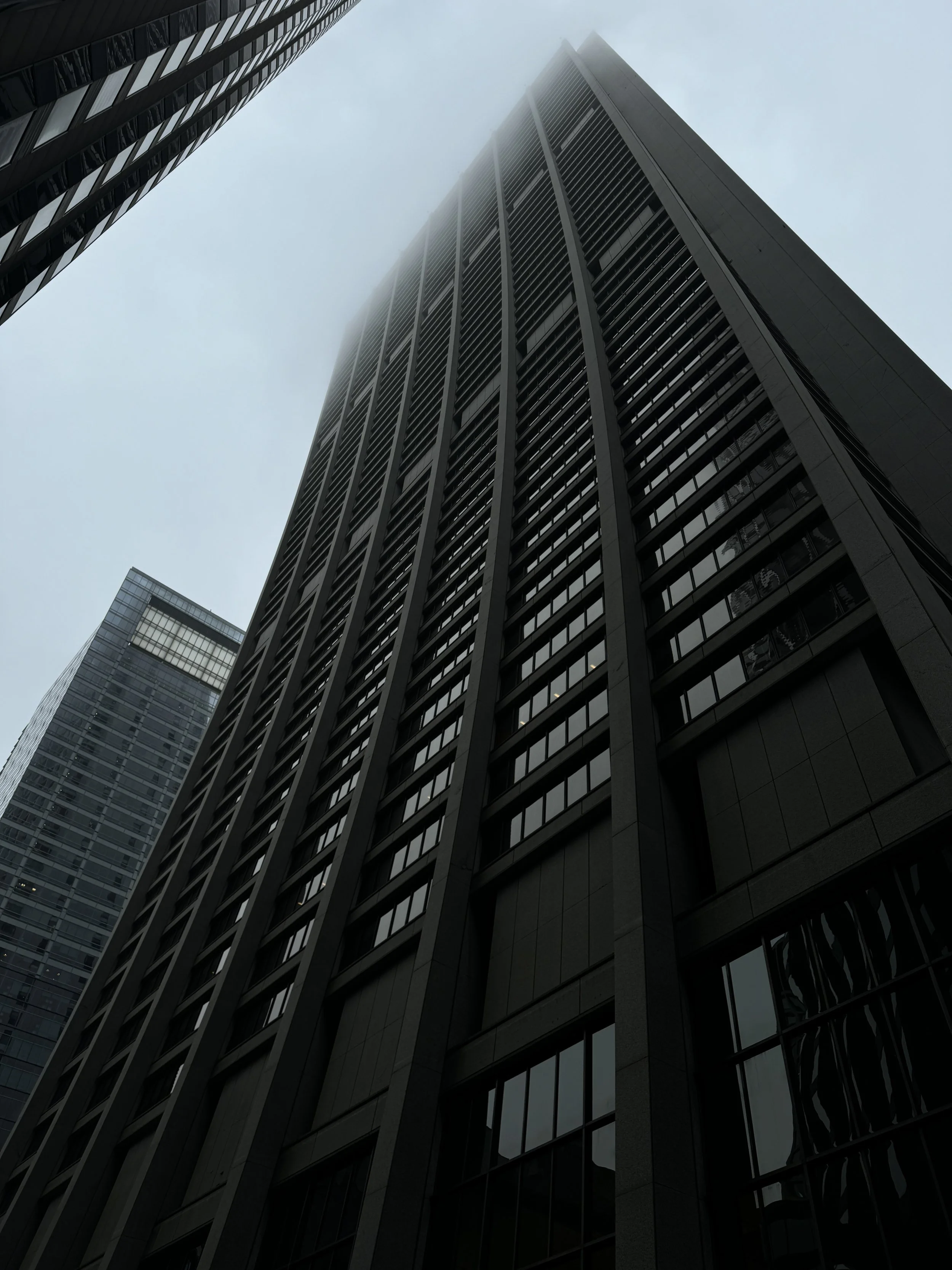 Low-angle view of modern skyscrapers with glass windows, one topped with fog or low-hanging clouds, in an urban setting.