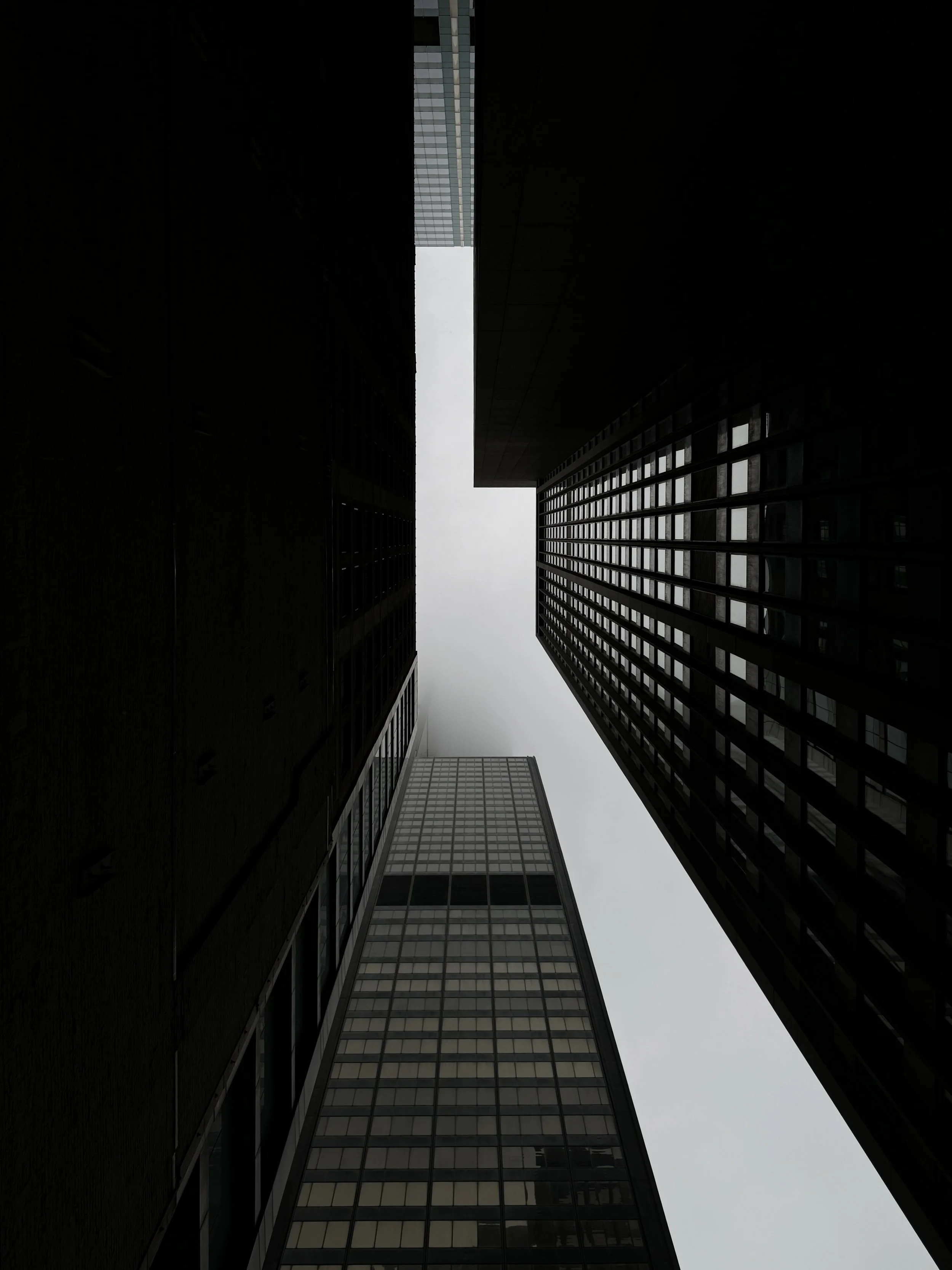 Looking upward between two tall skyscrapers with glass windows, with an overcast sky visible at the top.