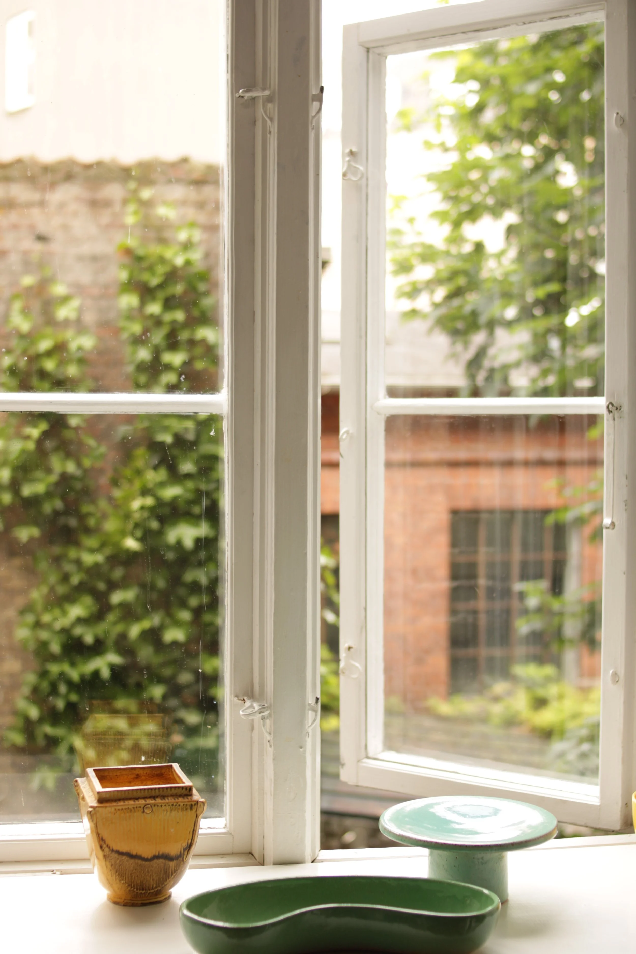 Open windows with a view of a brick building and green trees outside. Items on the windowsill include a yellow ceramic pot, green bowls, and a green ceramic stand.