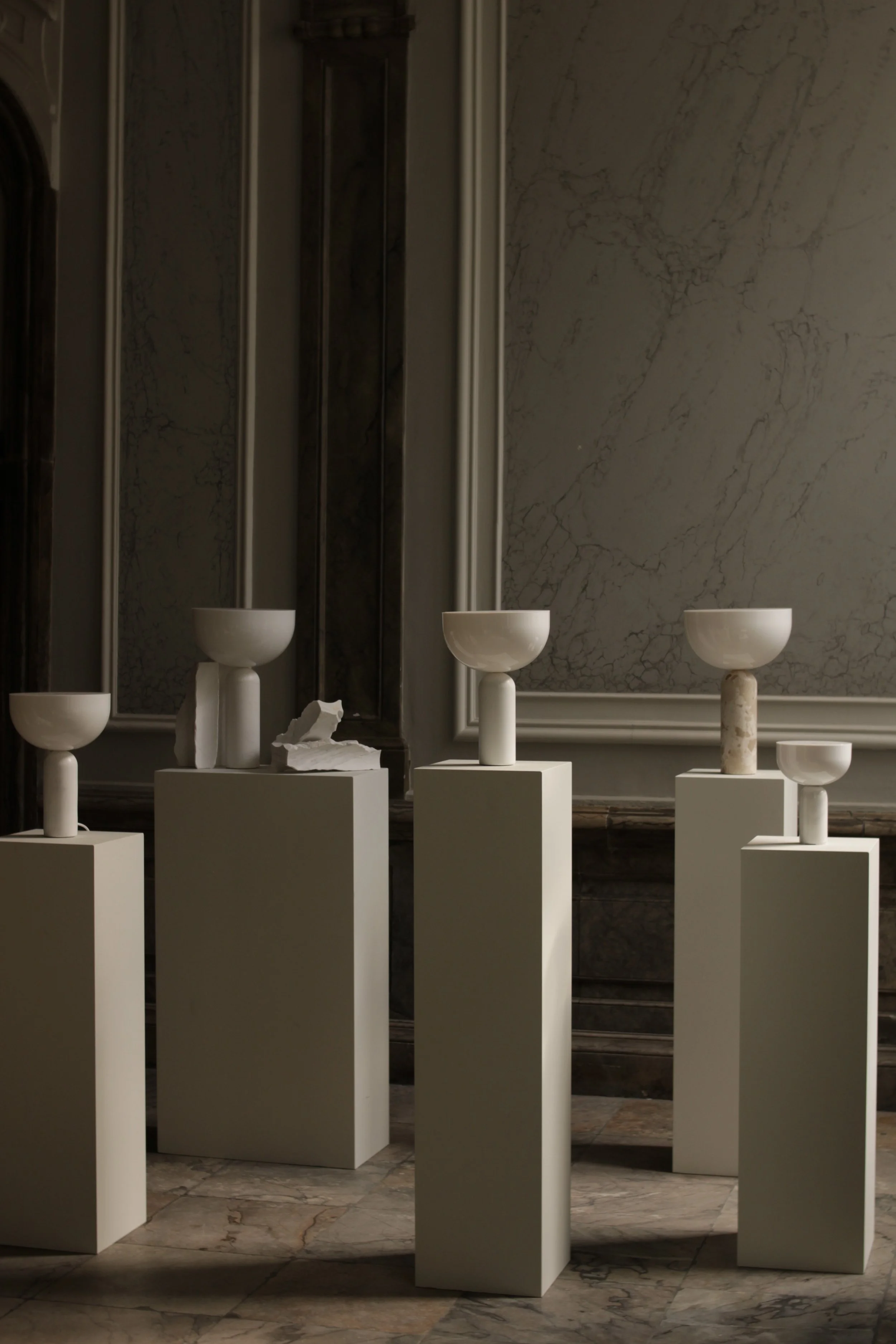 Art installation featuring white pedestal stands with ceramic bowls against a marble wall with textured gray and white veining in a gallery setting.