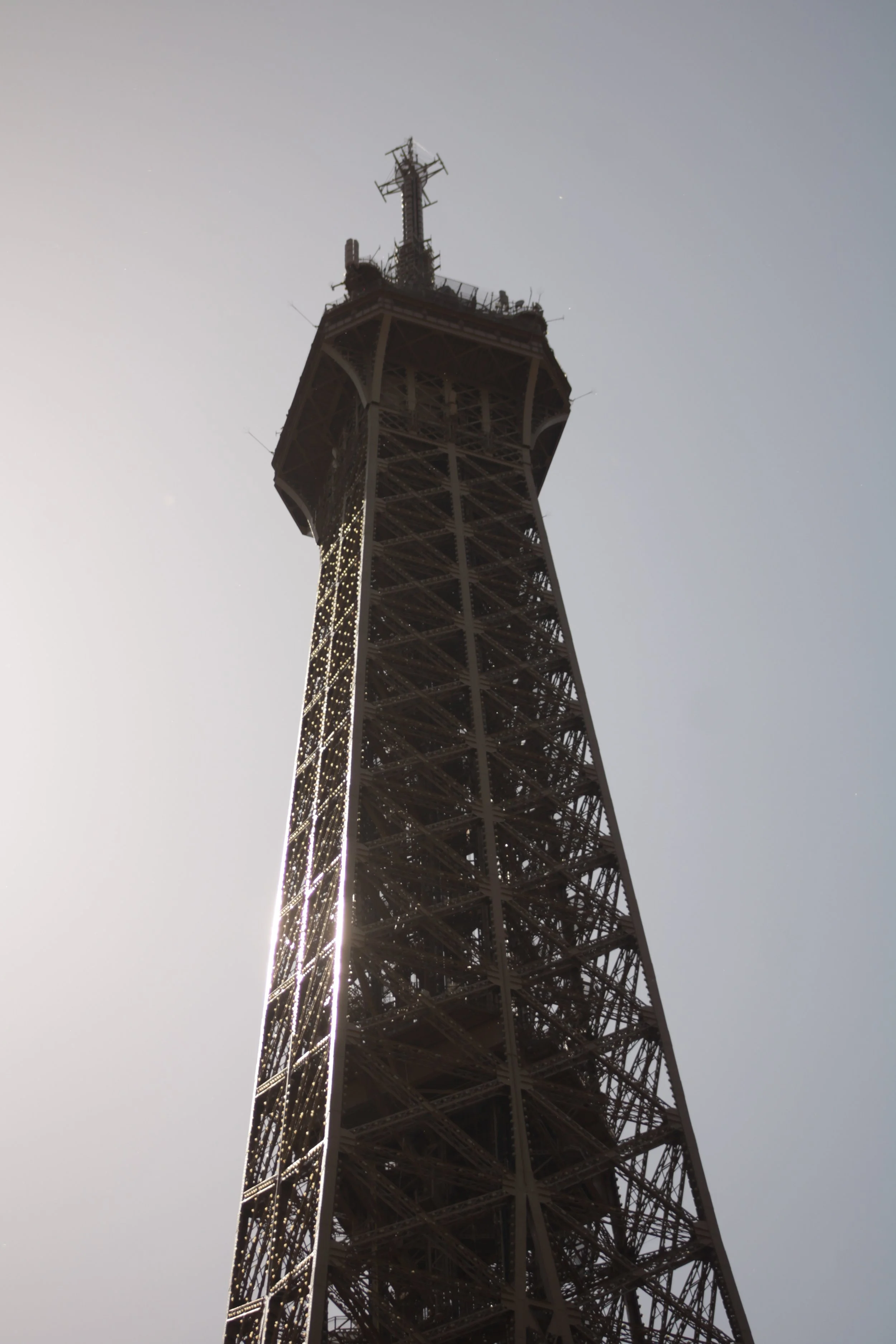 The Eiffel Tower in Paris viewed from below against a cloudy sky, with the top portion illuminated and showing signs of construction or maintenance.