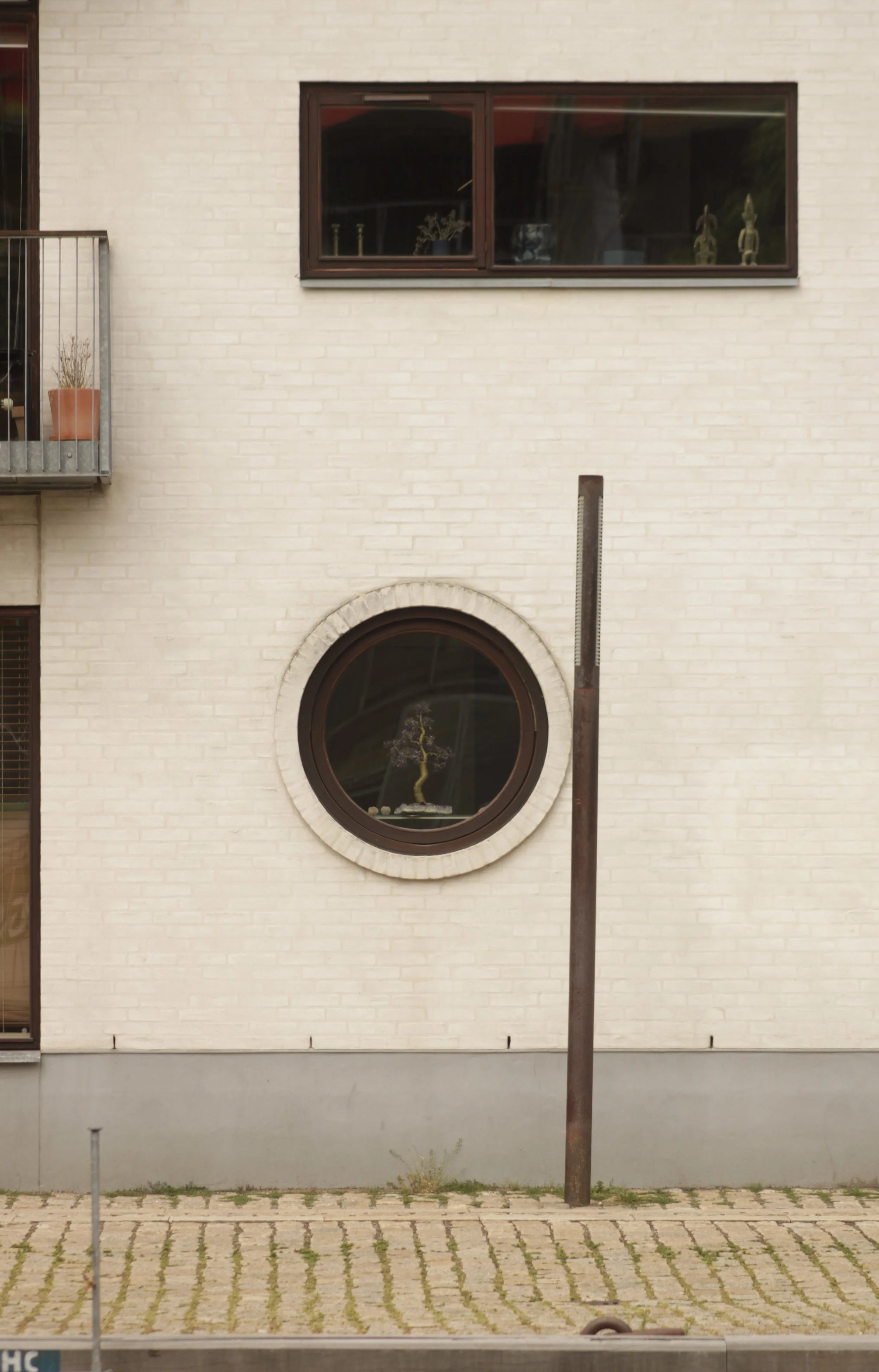 A beige brick building exterior with a round window displaying a small tree and decorative items inside, a rectangular window with plants and decor, and a tall rusted metal pole in front.