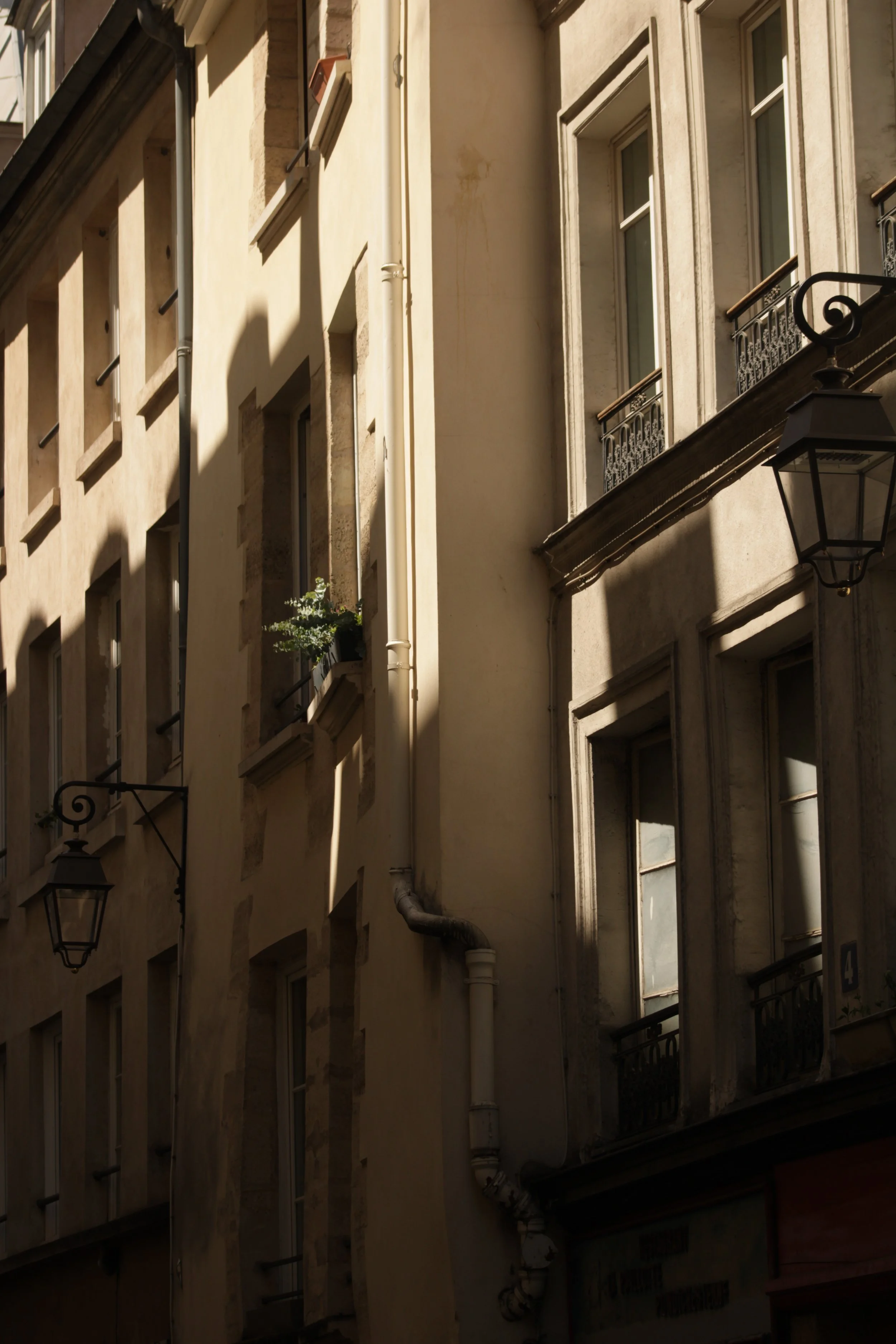 Close-up of a multi-story building facade with windows, wrought iron balconies, a street lamp, and shadows cast by neighboring structures.