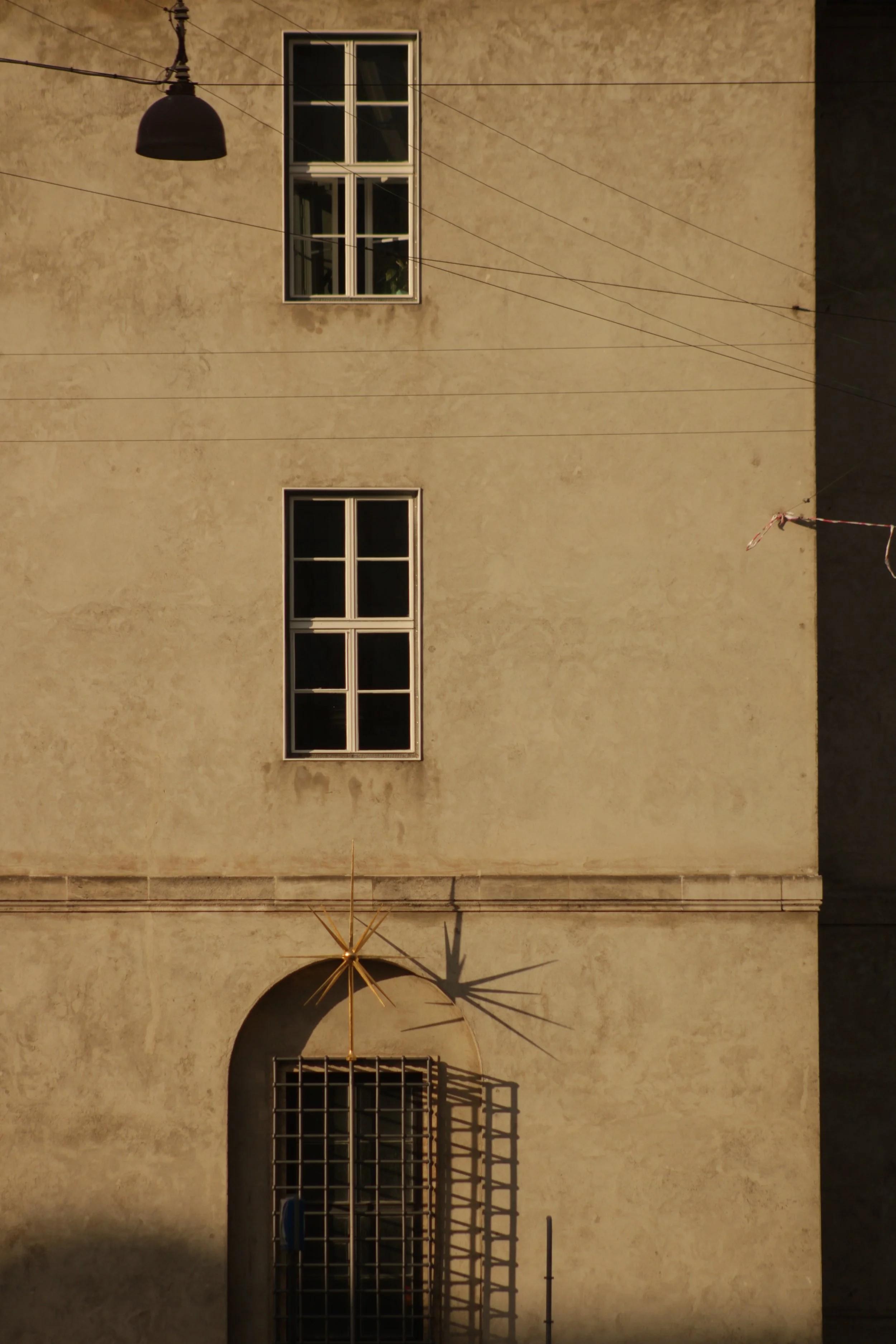 An exterior view of a beige building with four rectangular windows, a black street lamp, and some overhead wires. There is also a rolled-up awning with a metal framework below the lower window, and shadow cast by the wires and awning.