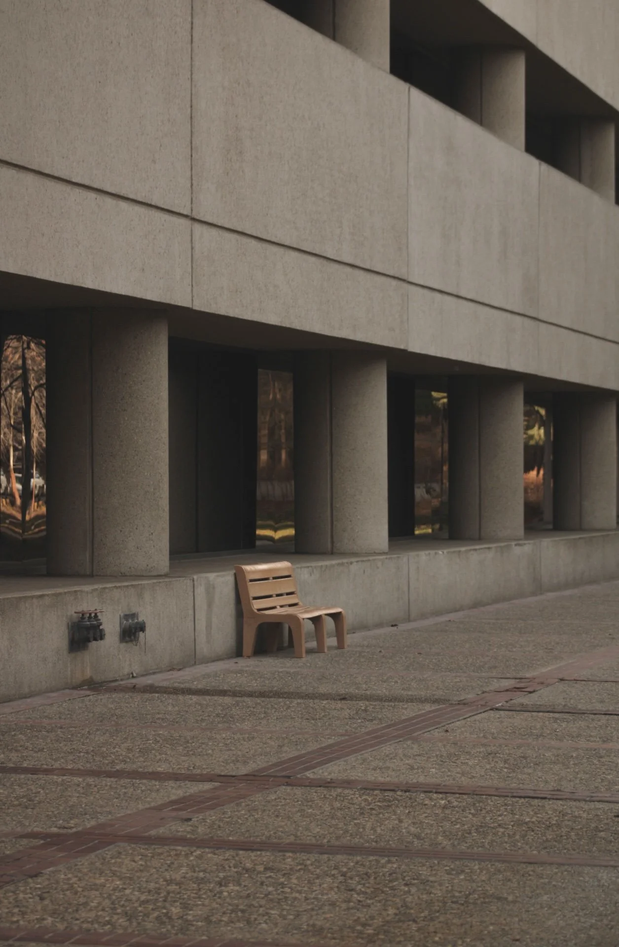 A concrete building with large vertical windows and a flat roof, with two fire hydrants on the sidewalk, a wooden bench, and trees visible through the windows.