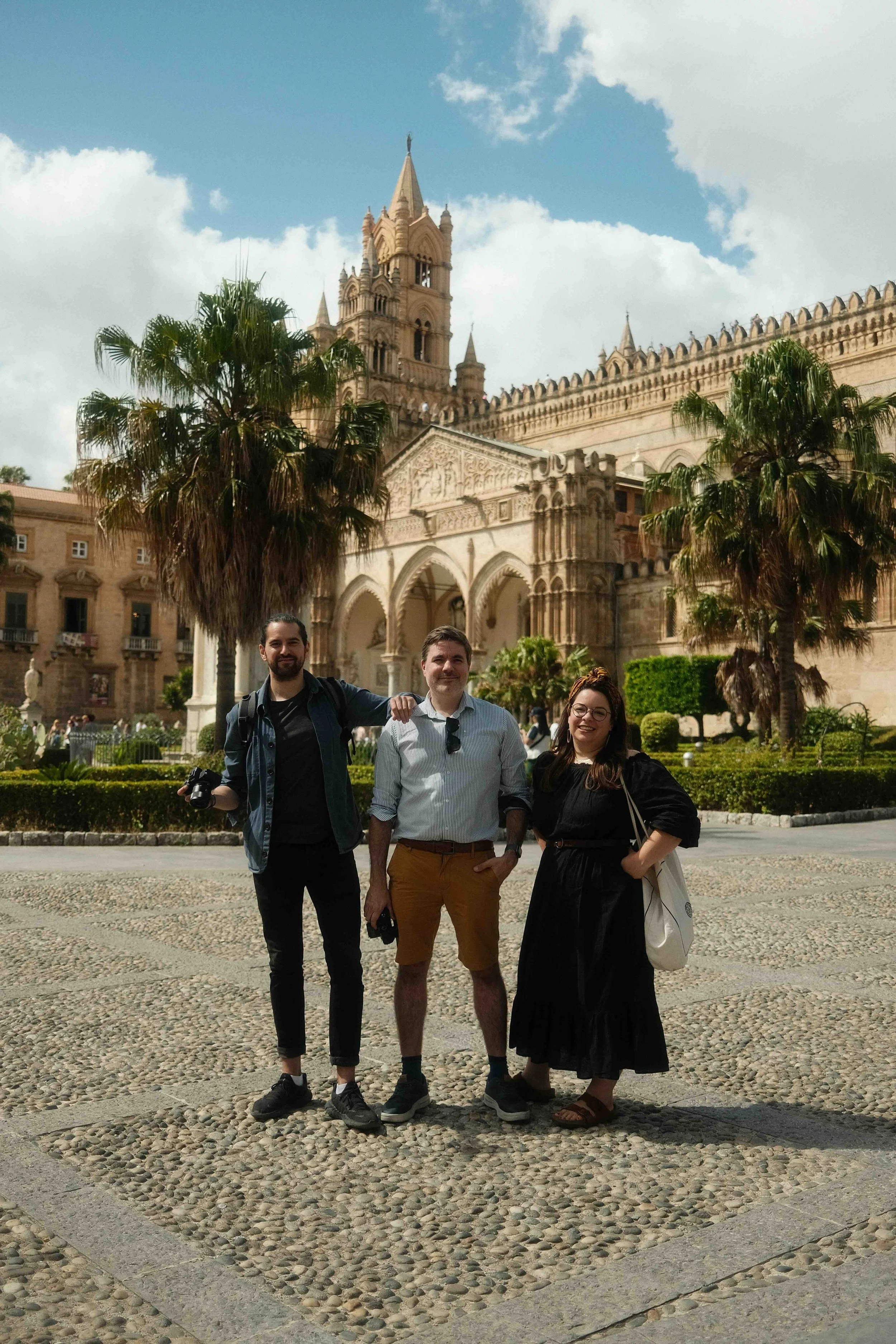 Three people standing in front of a historic cathedral building with palm trees and a cobblestone plaza.
