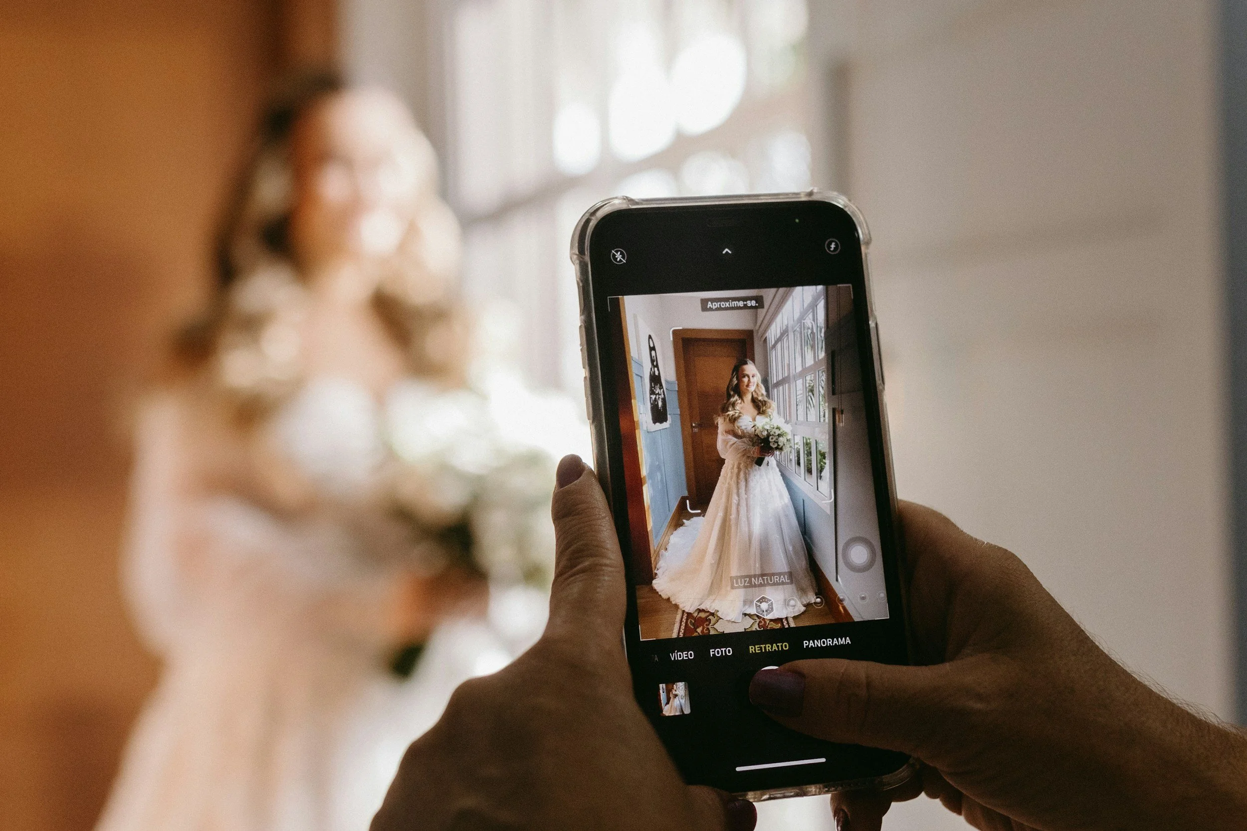 Person taking a photo of a bride holding a bouquet near a window with a smartphone.