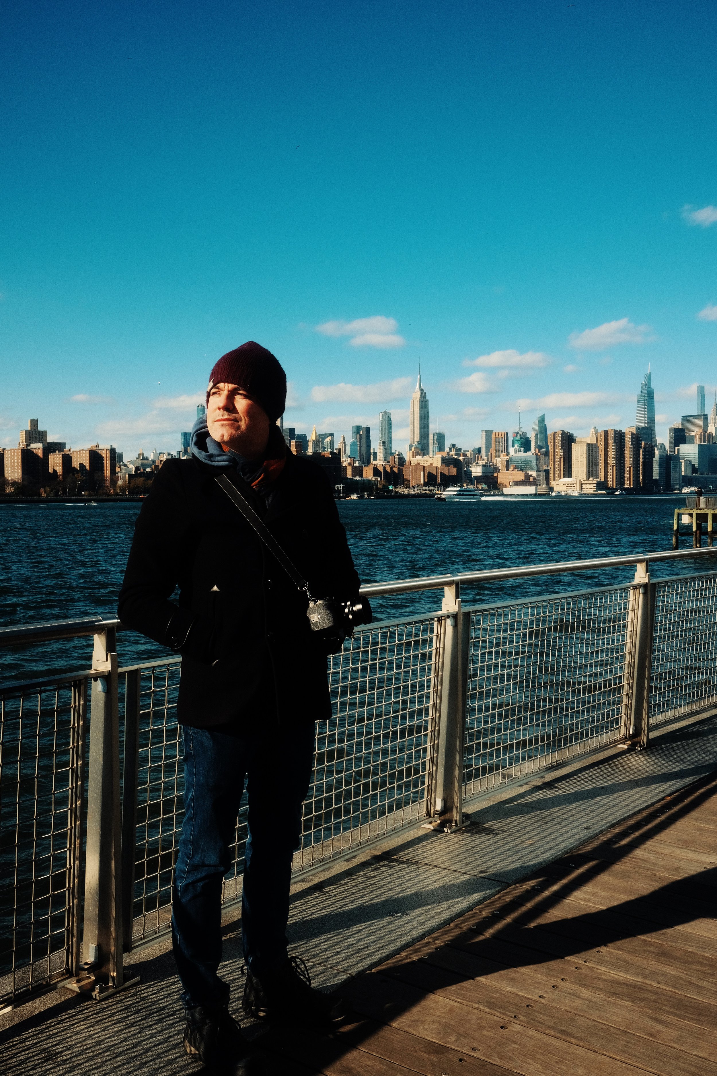 Person with a camera standing on a boardwalk overlooking a city skyline with a river in the foreground under a clear blue sky.