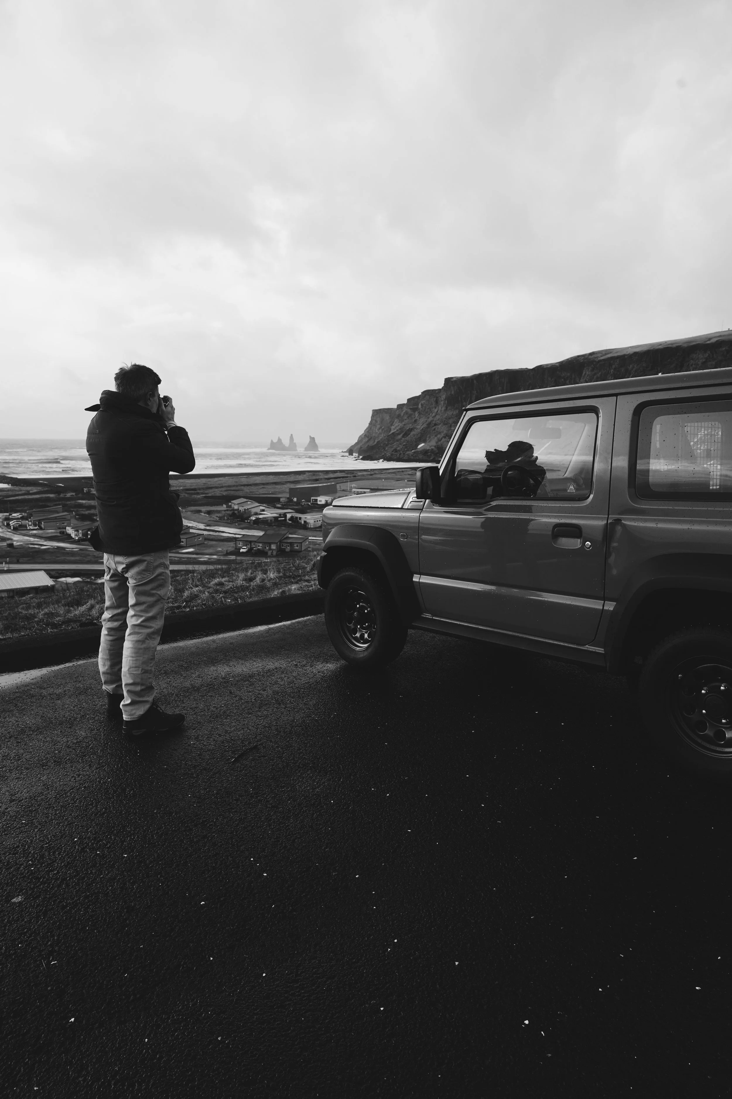 Man taking photo of ocean view with cliffs from roadside next to parked SUV, black and white image.