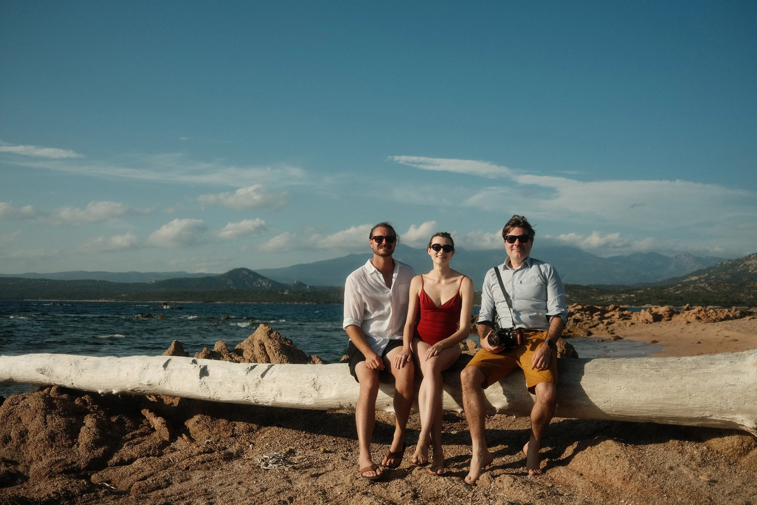 Three people sitting on a large driftwood log on a rocky beach with mountains and sea in the background.