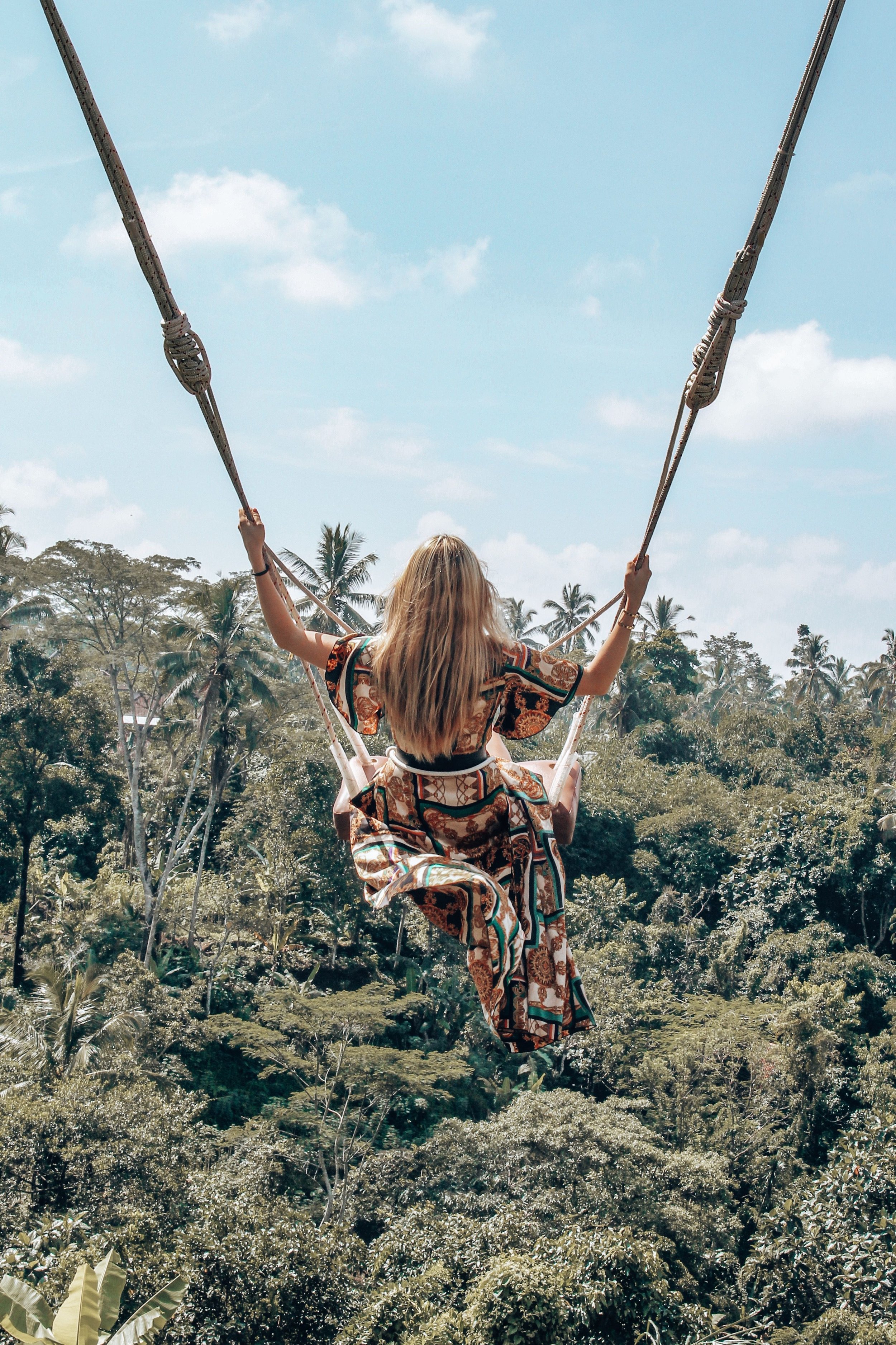 Mujer rubia en columpio sobre un paisaje tropical con árboles y cielo azul de fondo.