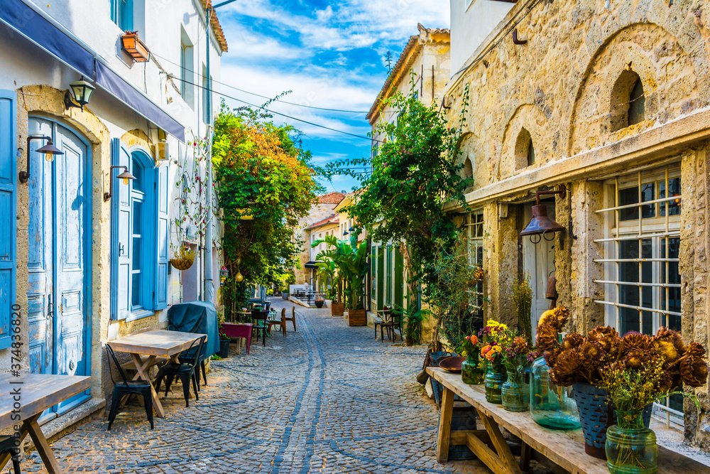 Calle empedrada estrecha con mesas al aire libre, puertas azules, edificios de piedra, plantas y flores en macetas.