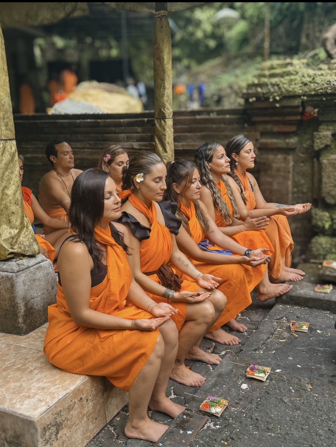 Grupo de personas sentadas en meditación, vestidas con túnicas naranjas, en un entorno al aire libre con estructura de piedra y naturaleza de fondo.