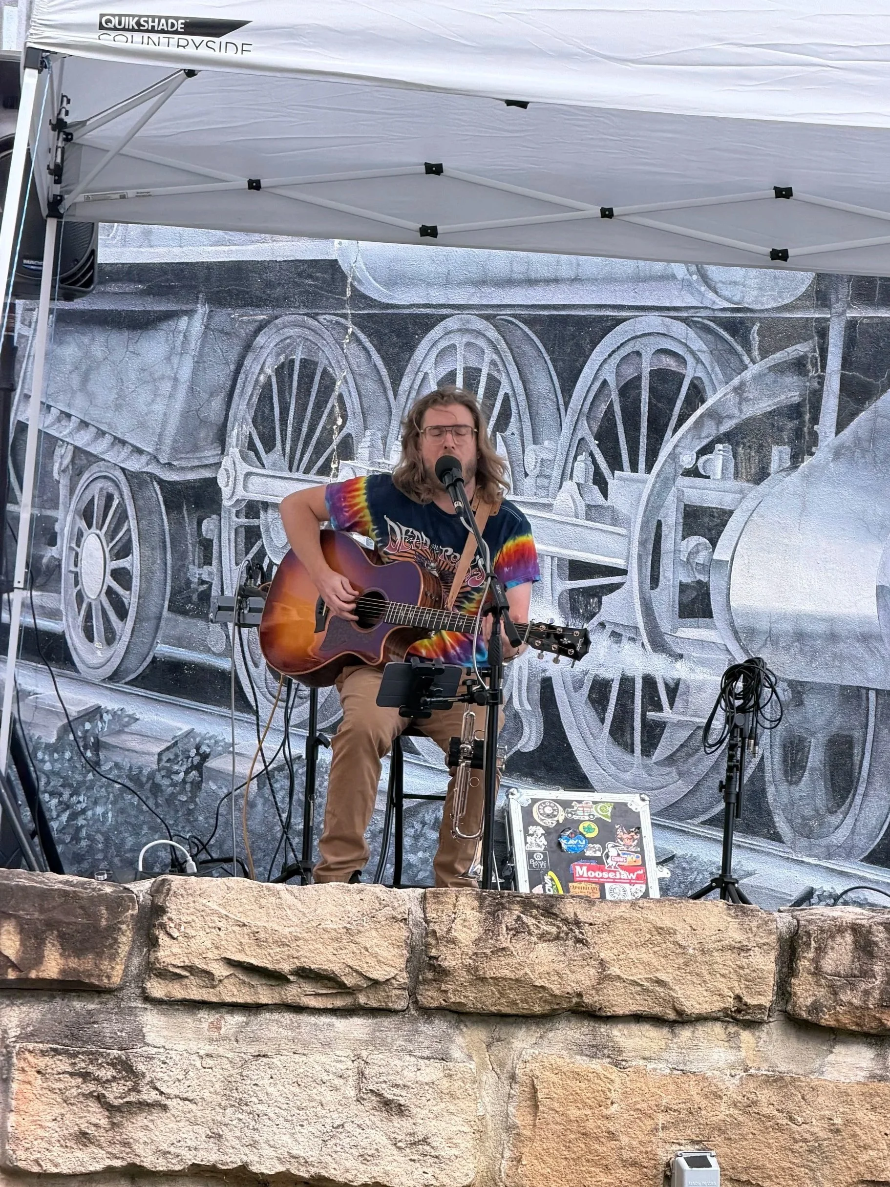 Person (Thomas Jude) playing guitar and singing on a stage with a monochrome train mural background, under a white canopy.