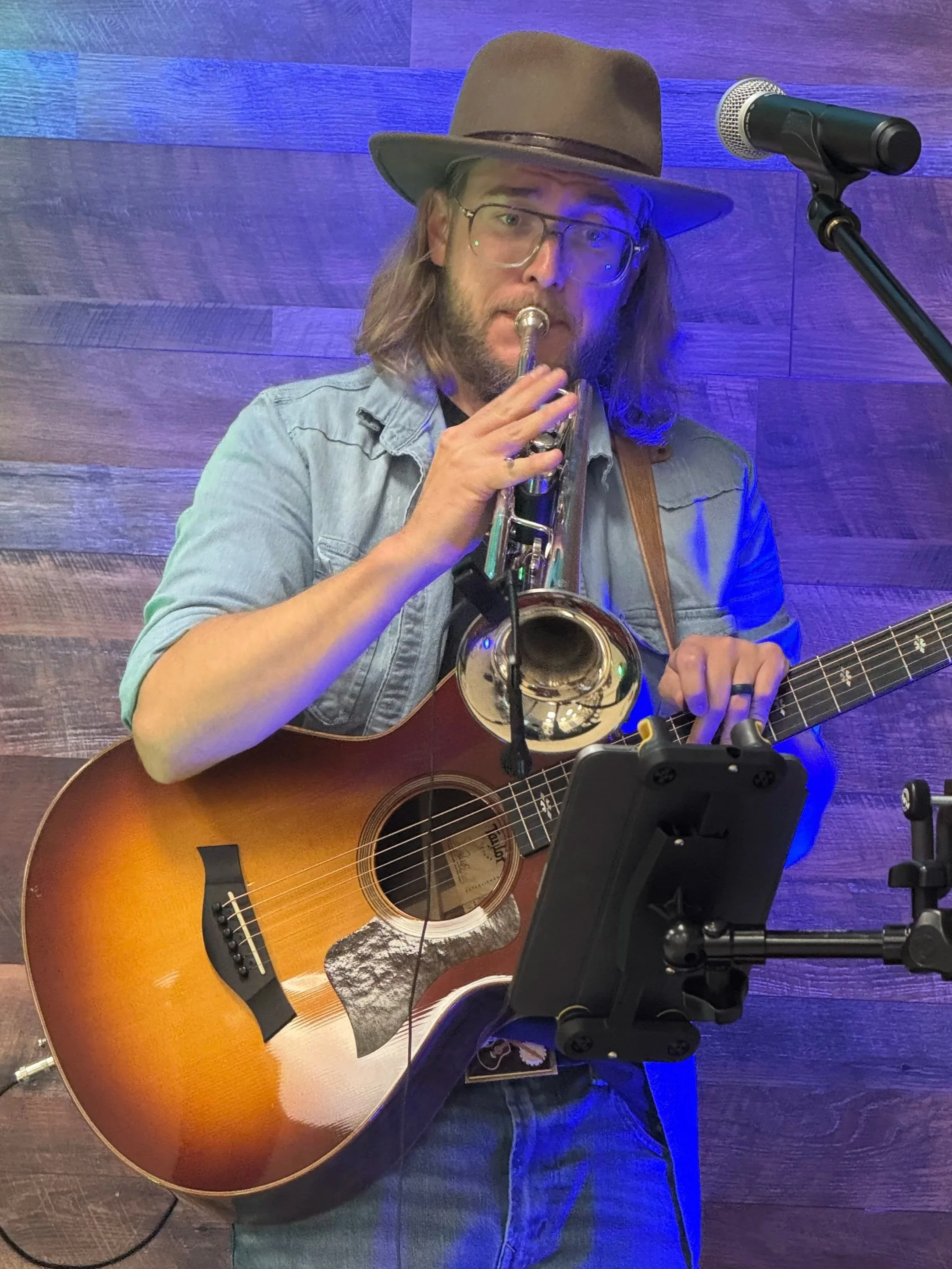 Musician (Thomas Jude) playing guitar and trumpet, wearing a wide-brimmed hat, glasses, and a denim shirt, standing in front of a wooden wall with a microphone nearby.