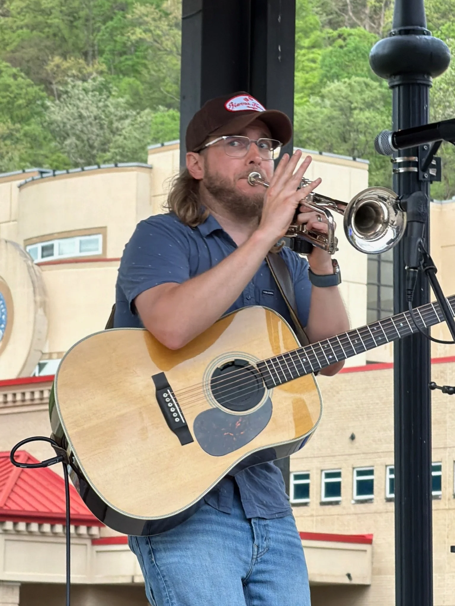 A man with glasses, a brown cap, and a blue shirt playing a trumpet and holding an acoustic guitar during an outdoor performance. A microphone is set up nearby, and in the background, there are green trees and a building with beige walls and multiple