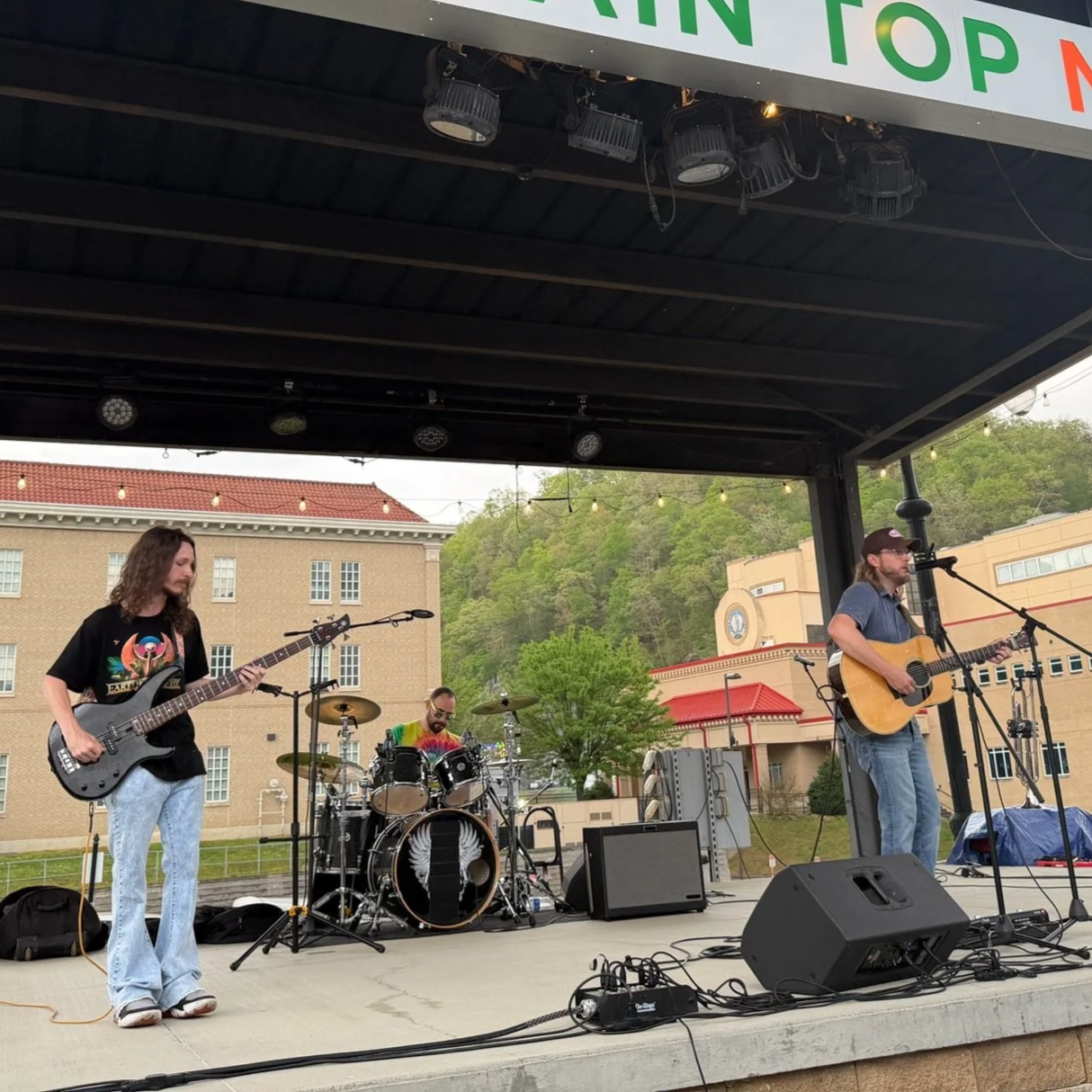 Three musicians performing on an outdoor stage with a building and trees in the background. One on bass guitar, one on drums, and one on acoustic guitar and singing into a microphone.