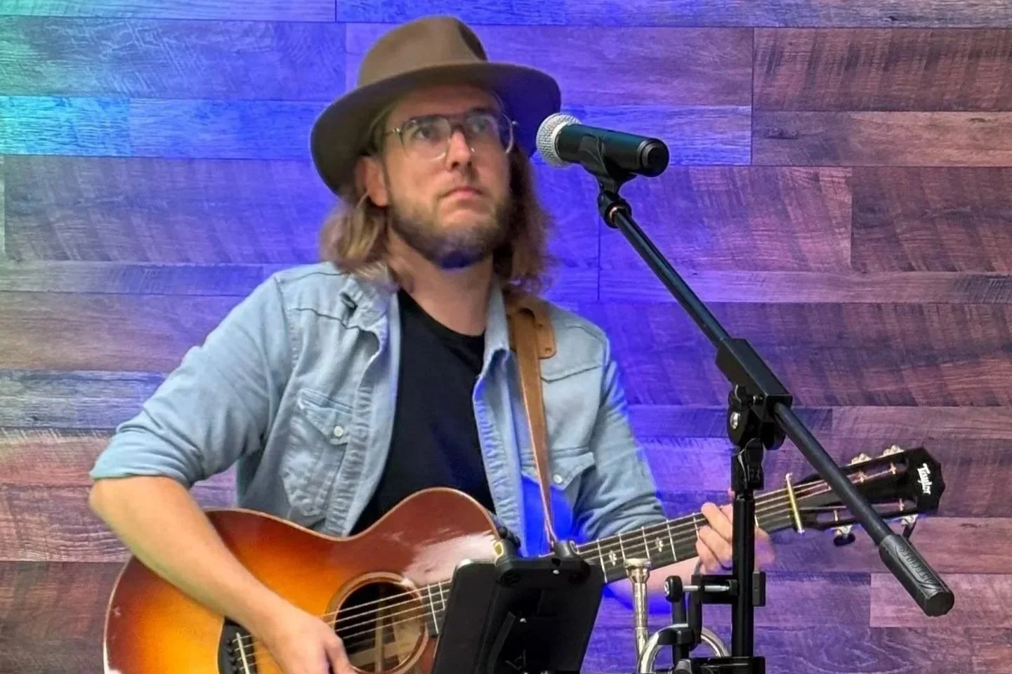 Man (Thomas Jude) with long hair and beard wearing glasses, a hat, and a denim shirt, playing an acoustic guitar near a microphone against a wooden wall background.