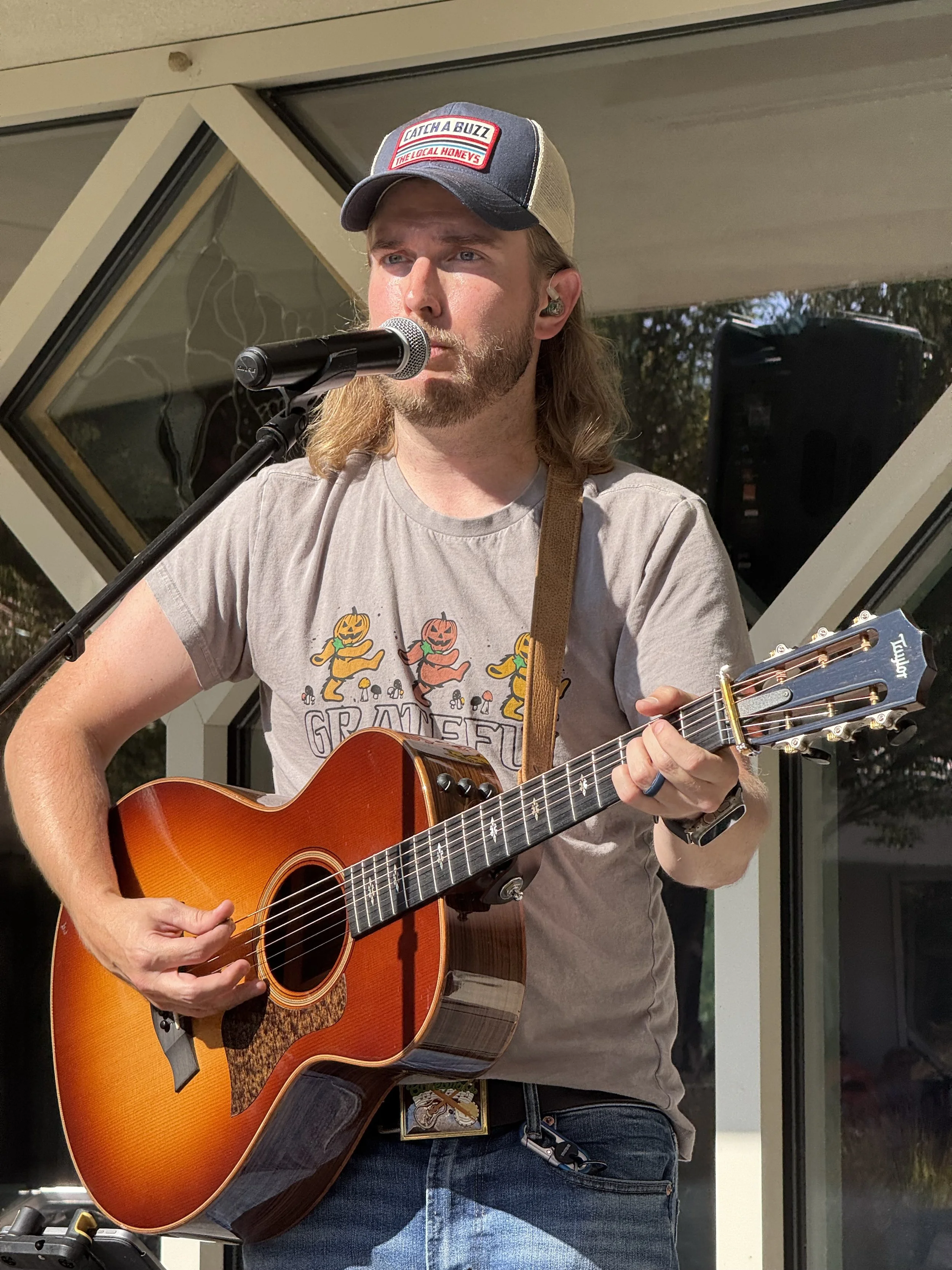 Thomas Jude singing into a microphone while playing an acoustic guitar. He is wearing a gray t-shirt, a baseball cap with red and white patches, and in-ear monitors. The background shows a modern building with large windows.