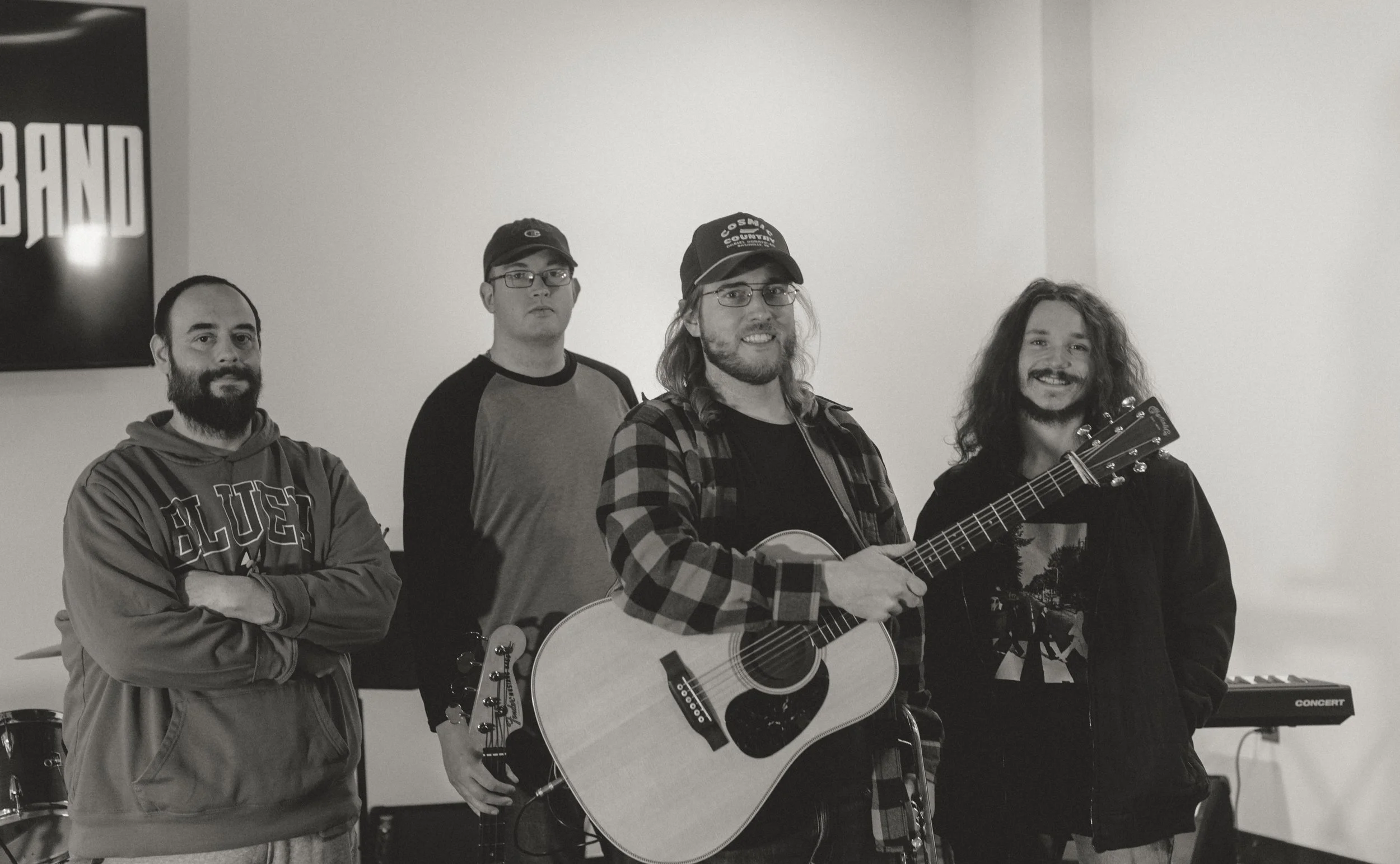 Four men standing indoors, one holding an acoustic guitar, with a keyboard in the background.