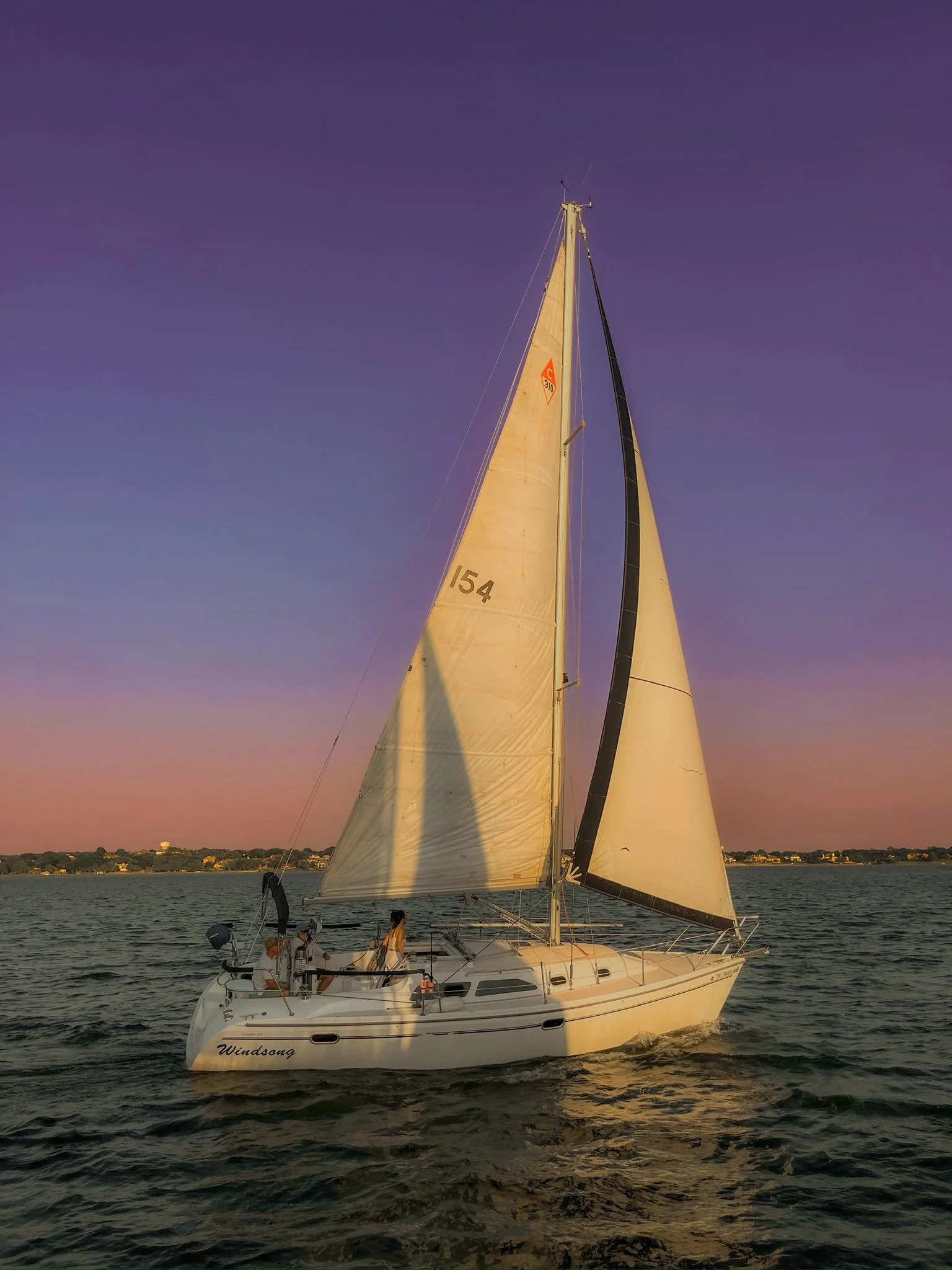 A sailboat named Windsong sailing on a body of water during sunset with a clear sky and distant shoreline.