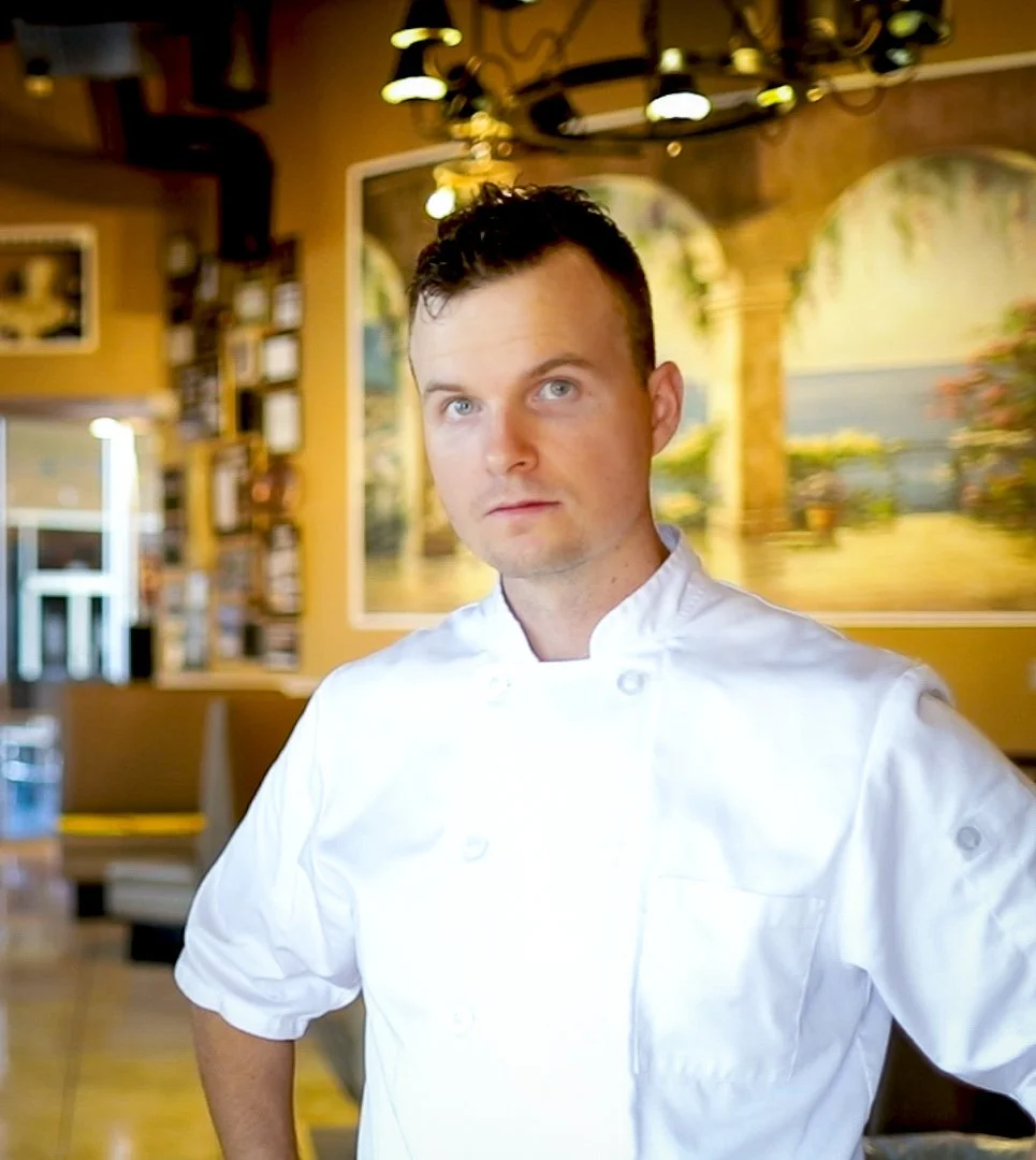 A male chef standing in a restaurant with a white chef's coat, looking at the camera. The restaurant's interior features warm colors, paintings, and decorative lighting.