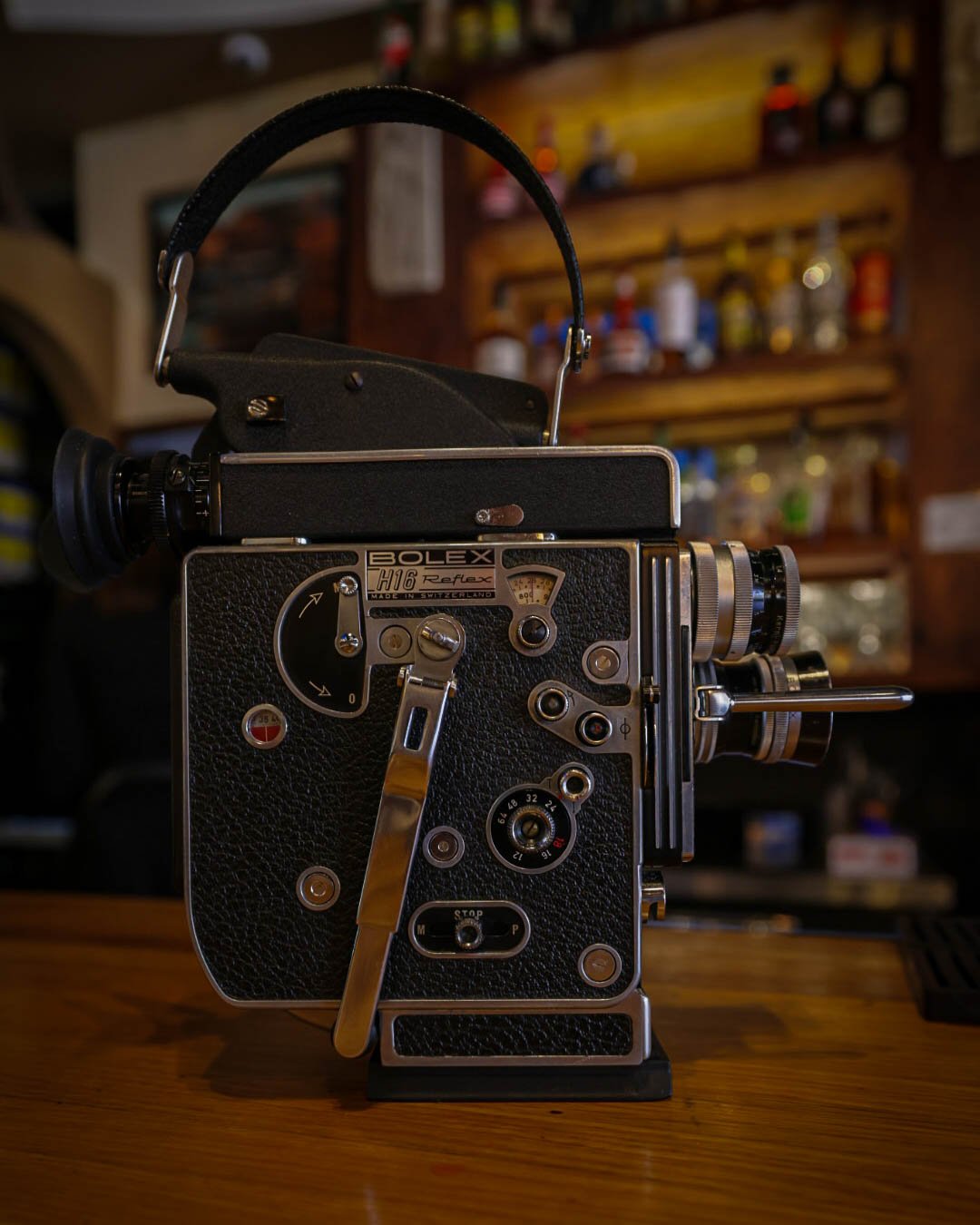 A vintage Bolex H16 Reflex 16mm film camera with black textured body, metal levers, knobs, and viewfinder, placed on a wooden surface with a blurred background of shelves and bottles.