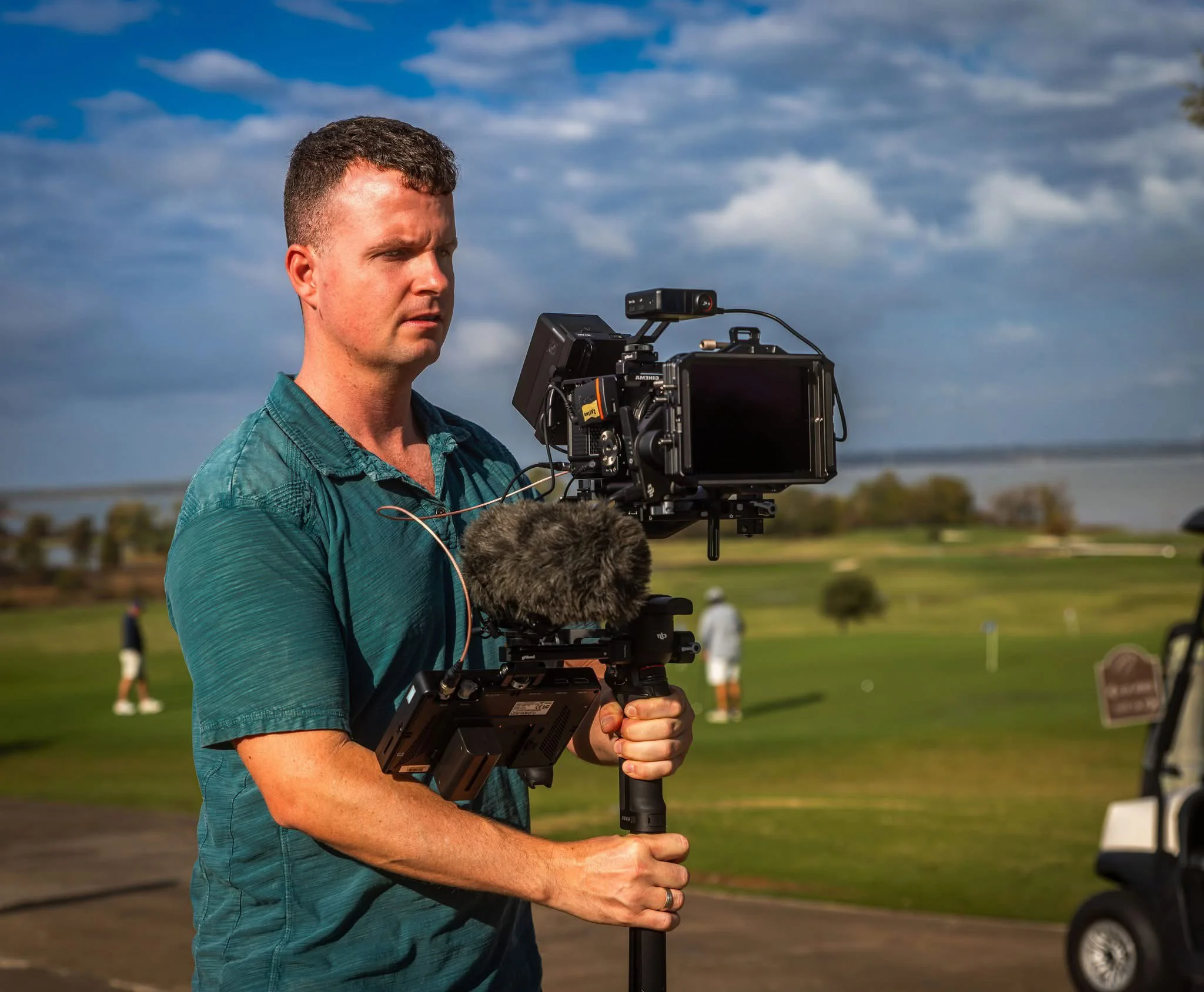 A man operating professional video equipment outdoors during the daytime, with a blue sky and clouds in the background, and some people visible in the distance on a grassy field.