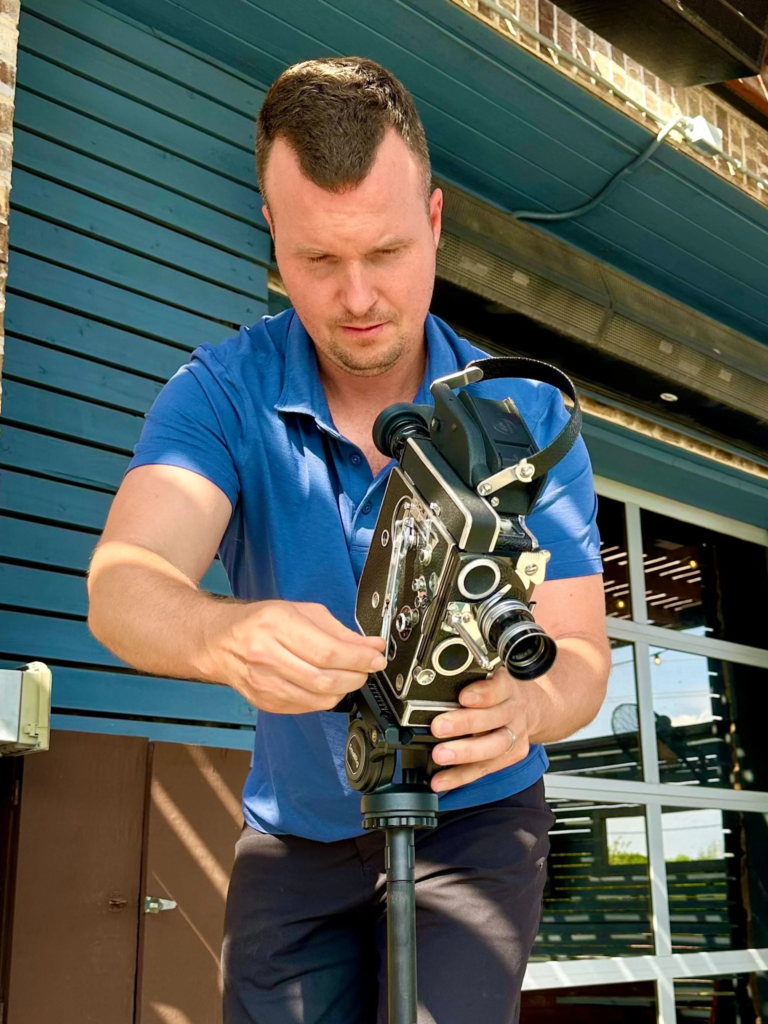 A man wearing a blue shirt and black pants is looking down at and adjusting a large, professional camera mounted on a tripod outside in front of a building with teal siding and brick accents.
