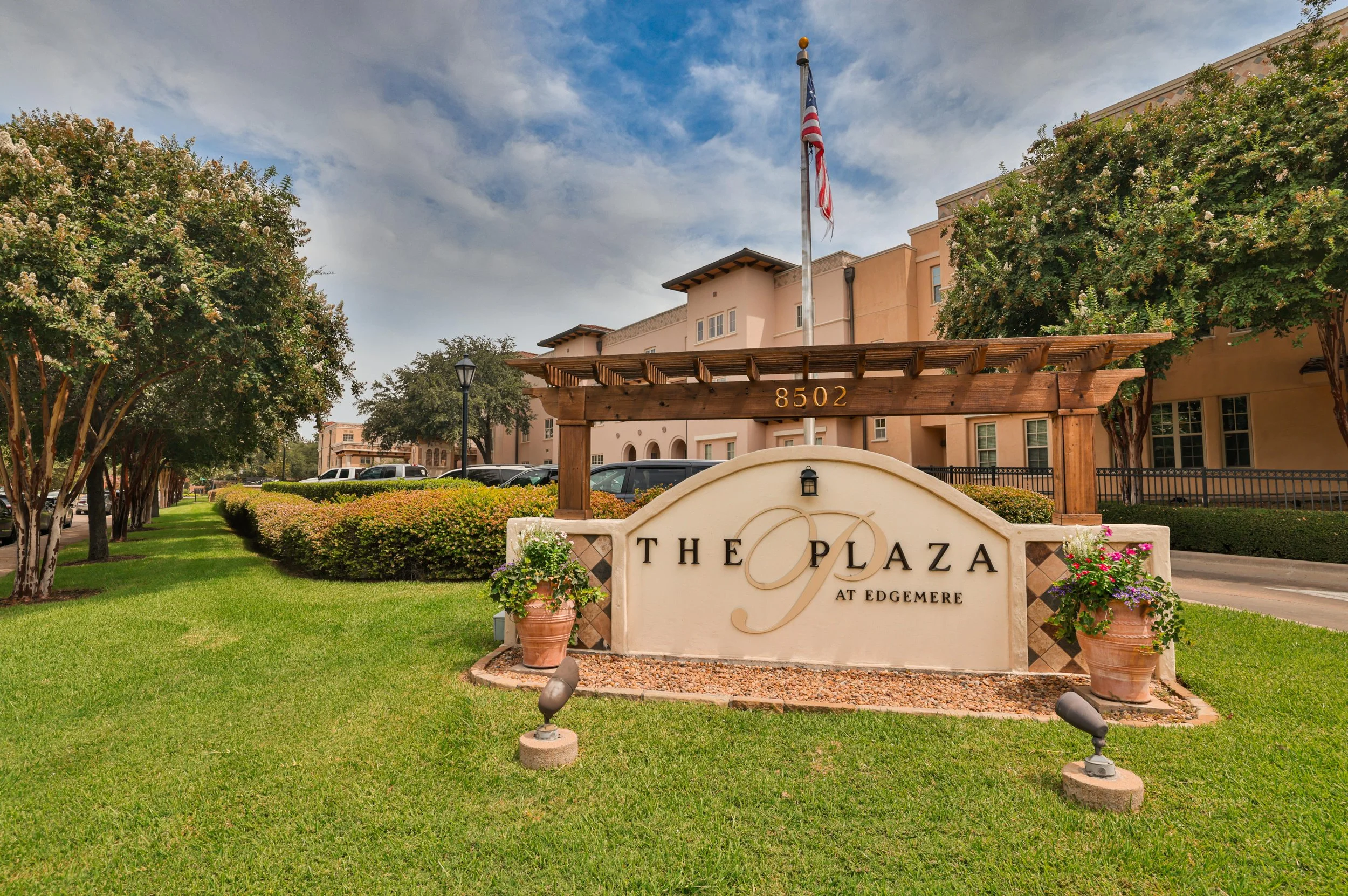 Entrance sign for The Plaza at Edgemere with potted flowers, surrounded by trees and parked cars in front of a beige residential building, with an American flag flying on a flagpole.