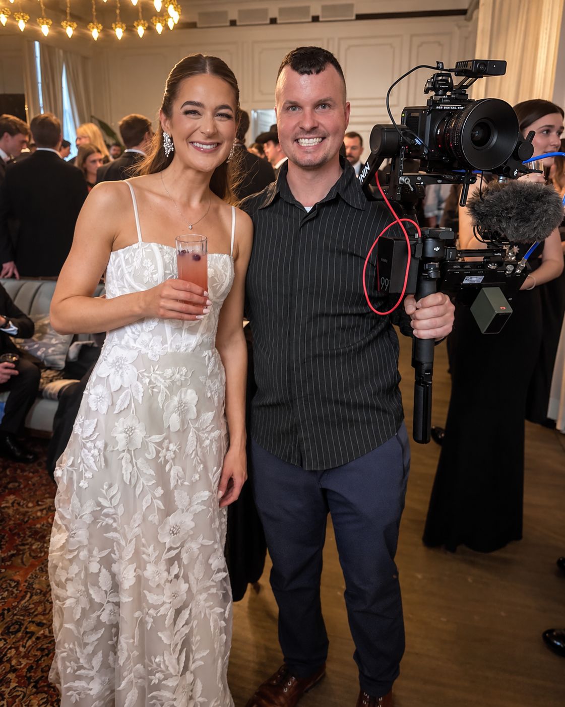 A woman in a white floral dress holding a pink cocktail standing next to a man in a black shirt holding a professional camera, at a formal event with many people dressed in formal attire in the background.