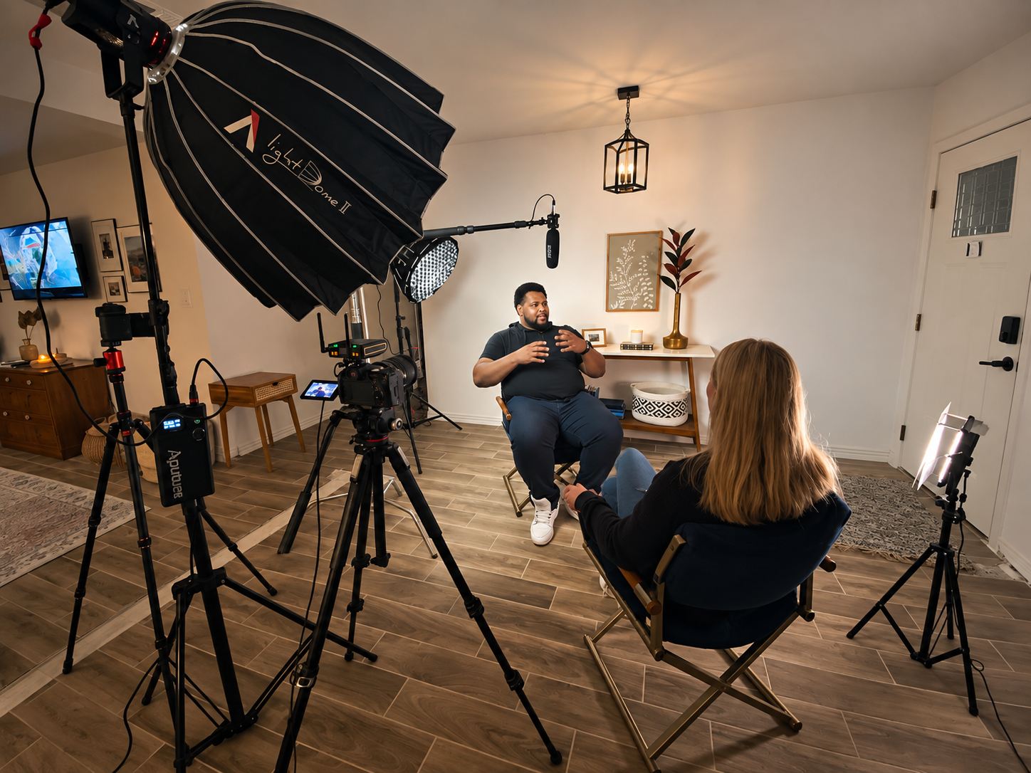 A man sits in a chair being interviewed by a woman in a small indoor studio space. There is professional camera equipment, lighting, and a microphone for recording.