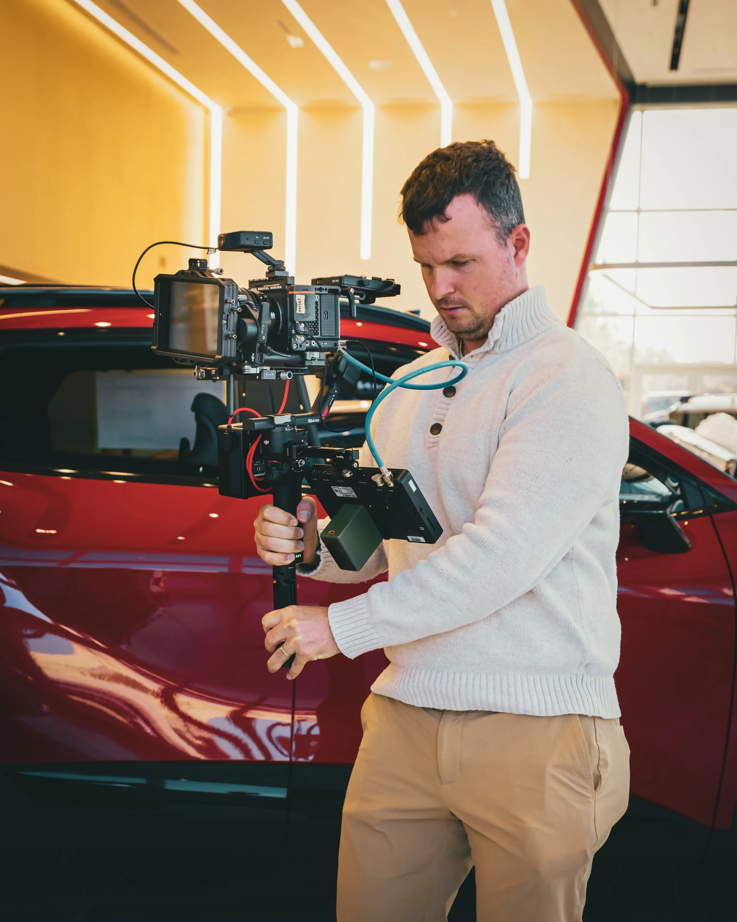 A man operating a professional camera mounted on a stabilizer inside a car dealership, with a shiny red car behind him and modern lighting overhead.