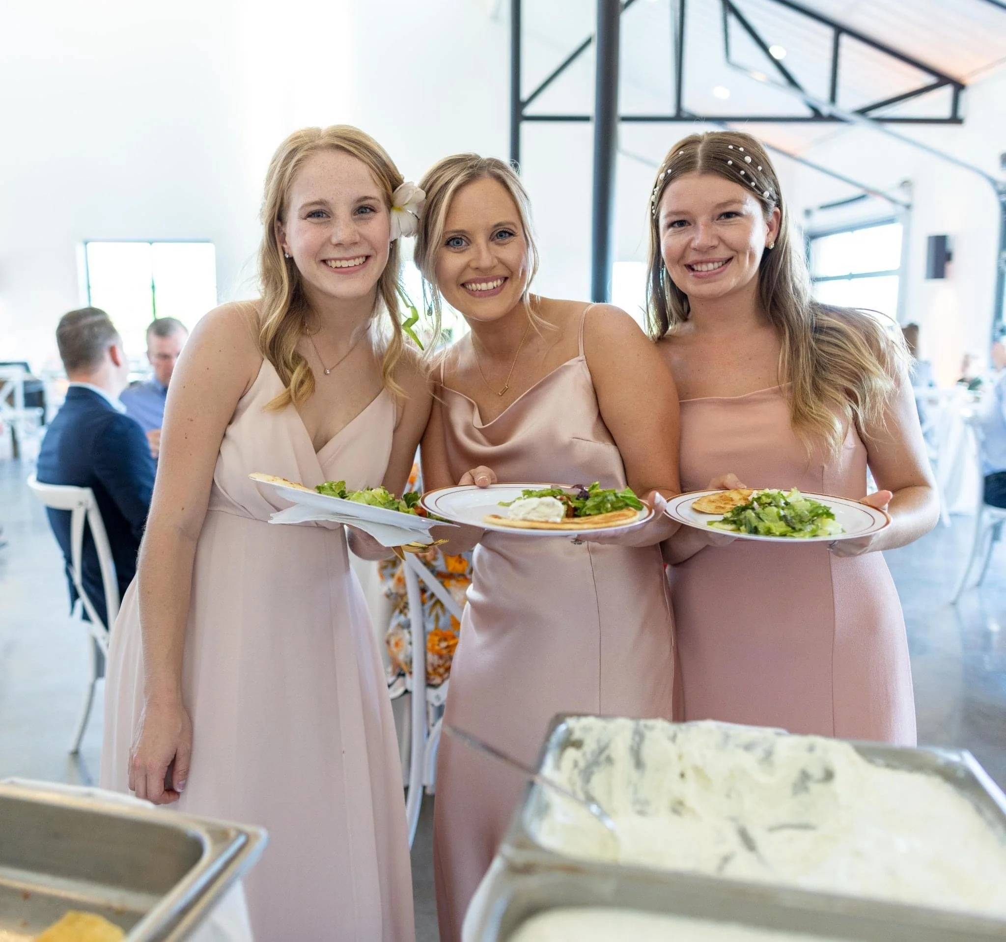 Three women dressed in pink dresses standing closely together and smiling in a bright indoor setting. They are holding plates with salads and appetizers, likely at a social gathering or celebration.