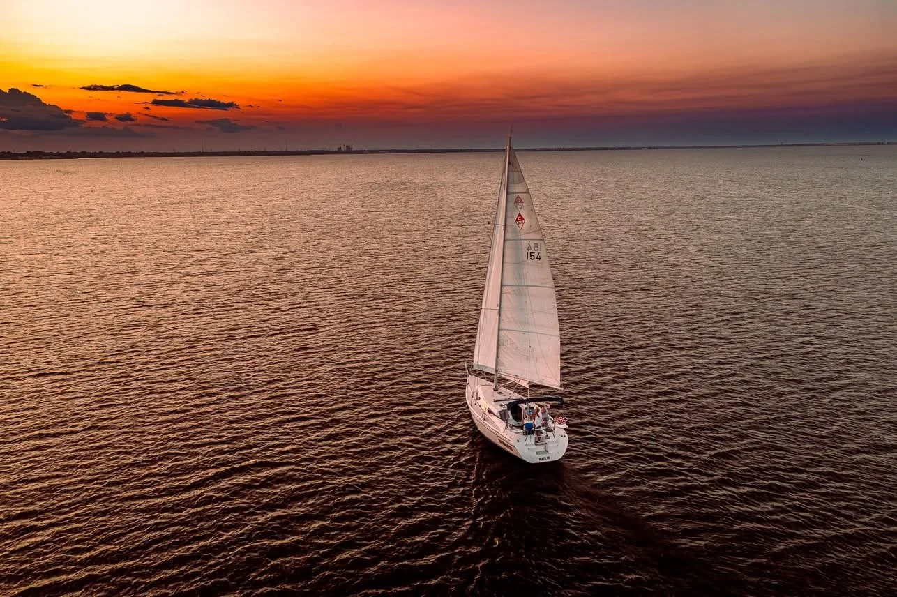 A sailboat with white sails sailing on a calm body of water during sunset with a colorful sky.