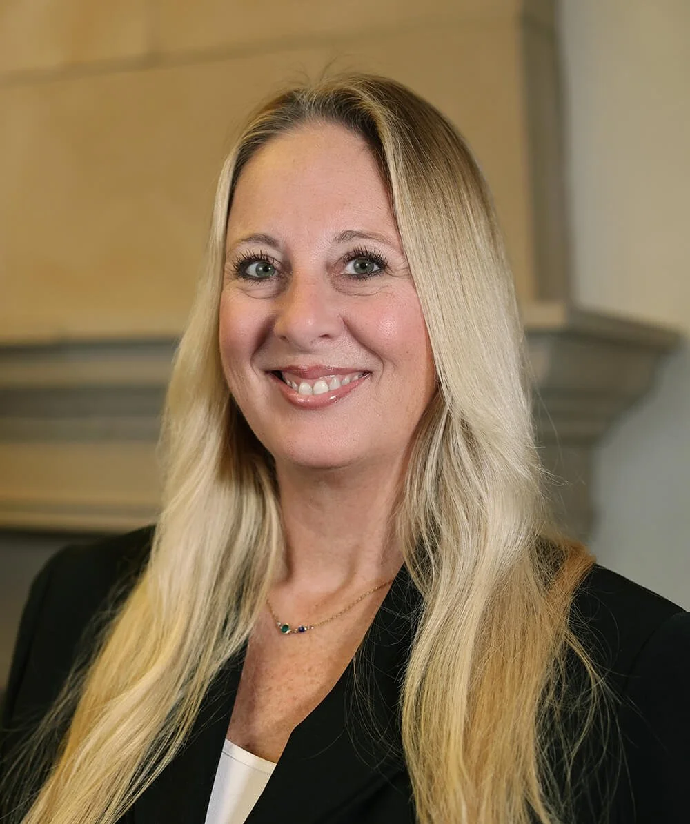 A smiling woman with long blonde hair, wearing a black blazer and a delicate necklace, is posing indoors against a neutral background.