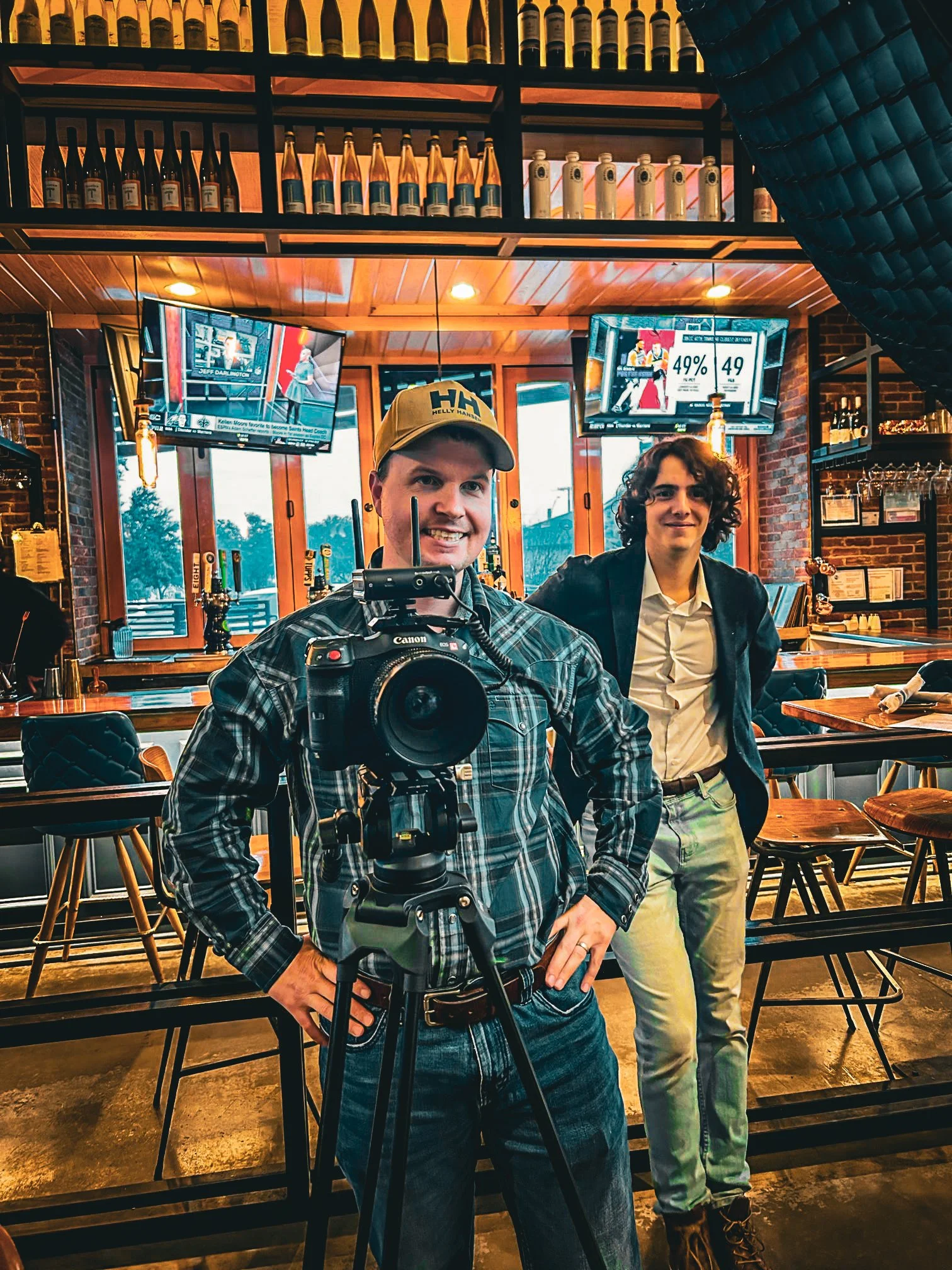 Two men in a bar or restaurant, one with a camera on a tripod taking a picture, and the other standing beside him. The background shows televisions and shelves with bottles.
