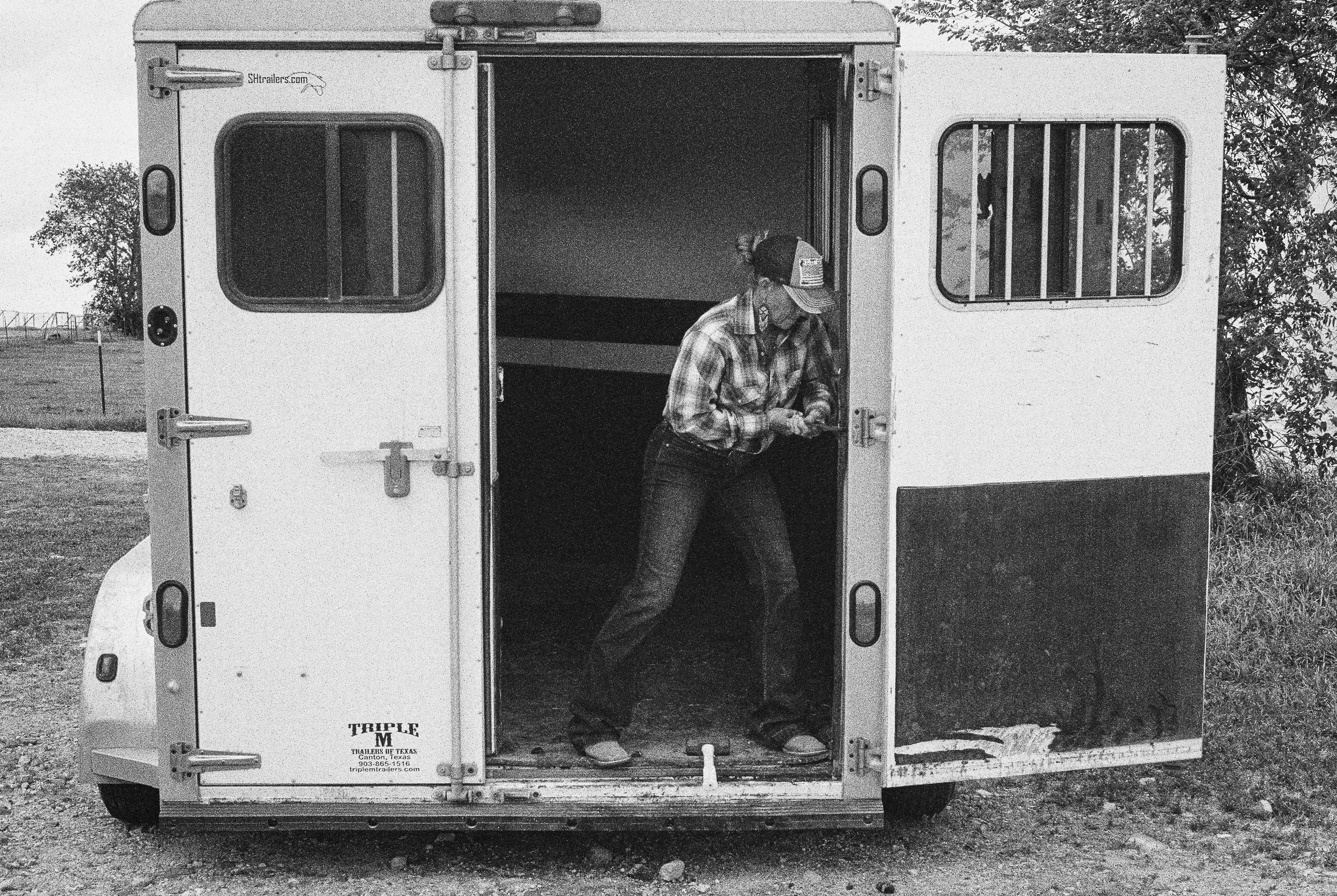 A man in a plaid shirt and baseball cap standing inside the open back of a white trailer truck, looking down. The trailer has barred windows on the sides and some writing on the back door, including a website and location information.