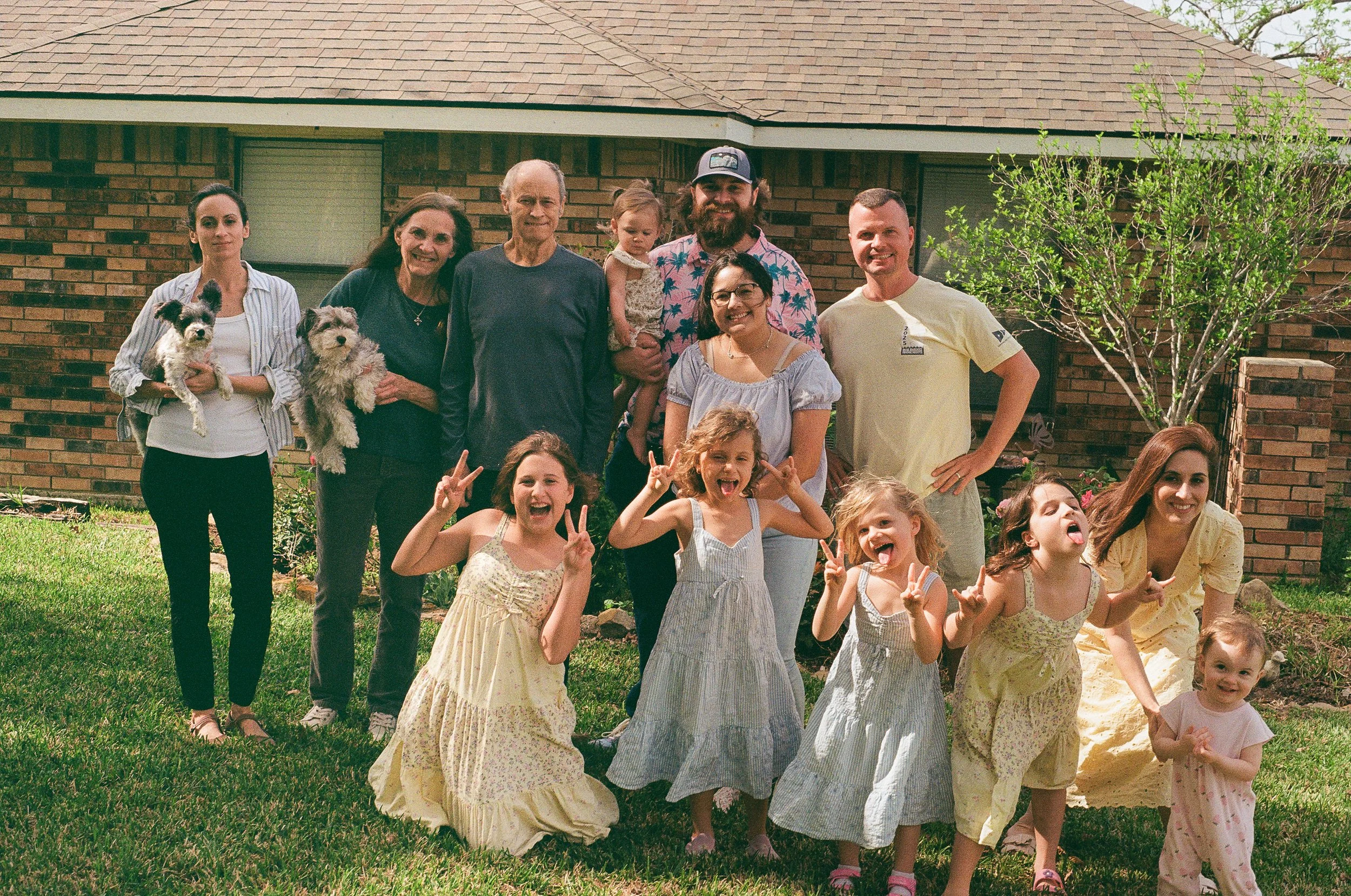 A group of children and adults gathered outdoors in front of a brick house, posing for a photo. The children are making peace signs and smiling, while some adults are holding puppies and standing together.