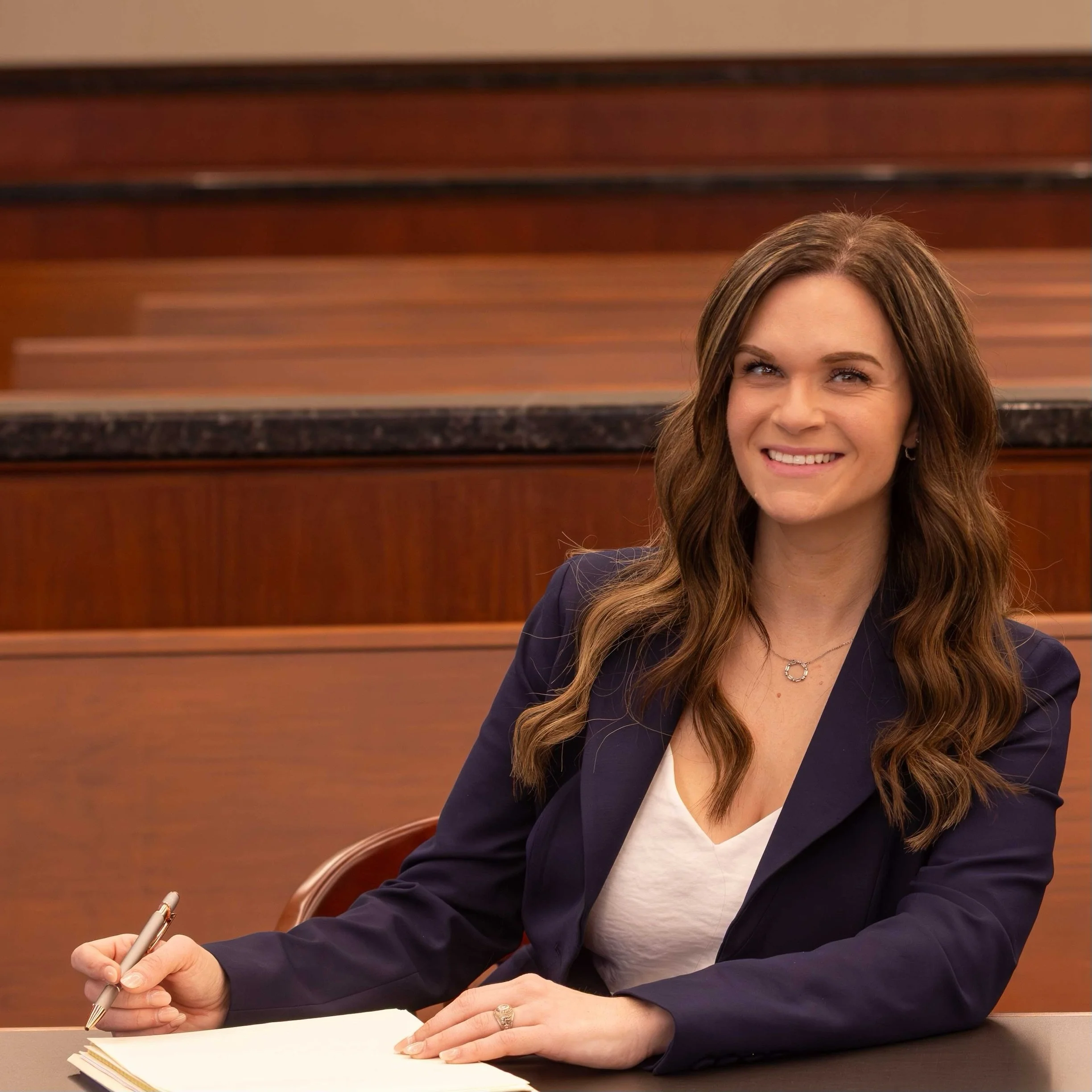 A woman with long brown hair, wearing a dark blazer and a white top, sitting at a table with a pen in her right hand, holding a stack of papers or a notebook. The background contains wooden wall panels.