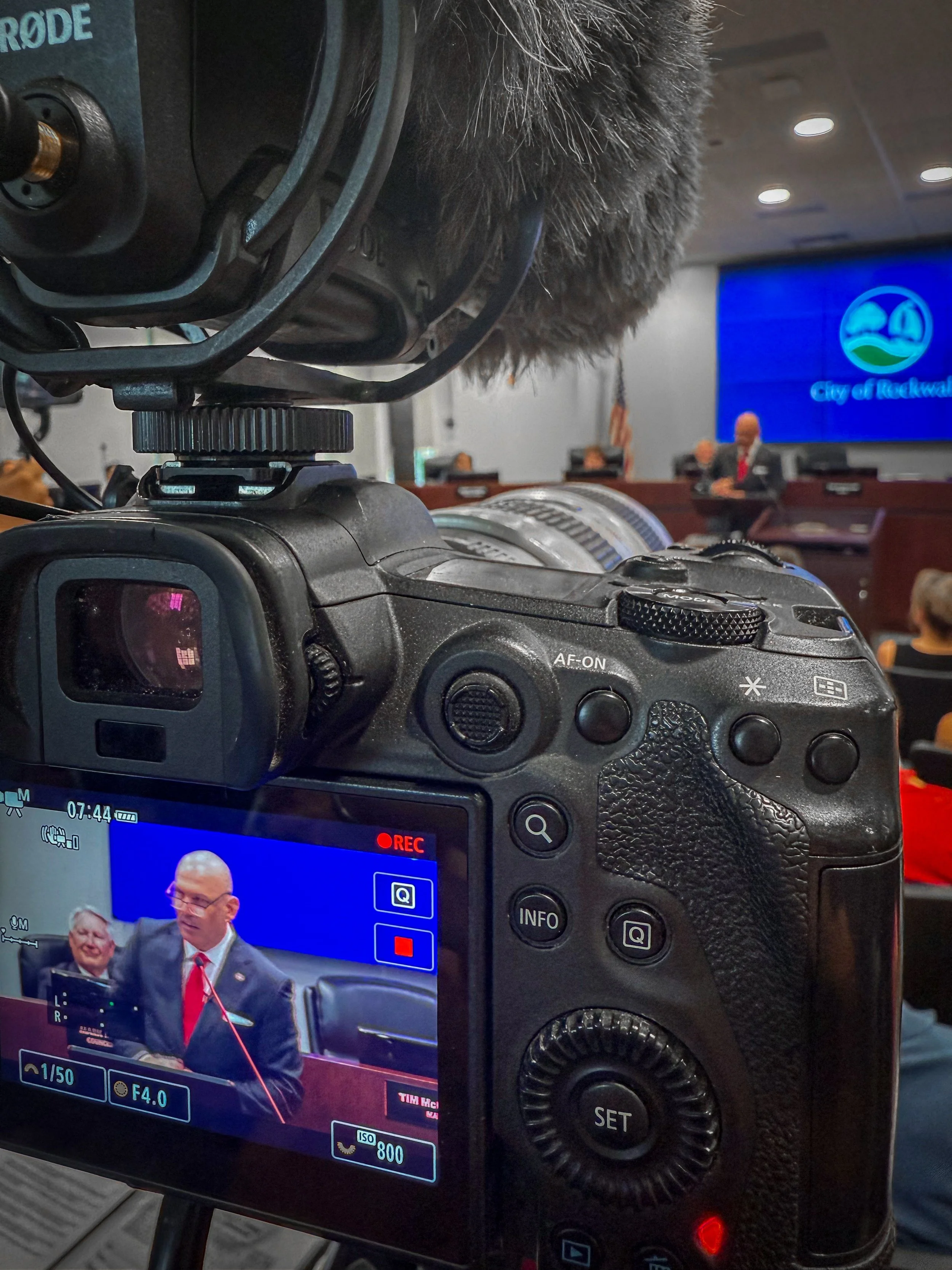Close-up of a professional DSLR camera with a display screen showing a live image of a conference room with people sitting at a panel. The camera is tilted, capturing part of its controls and a microphone with a furry windscreen. In the background, there is a digital screen displaying the logo of the City of Rockwall and some attendees.