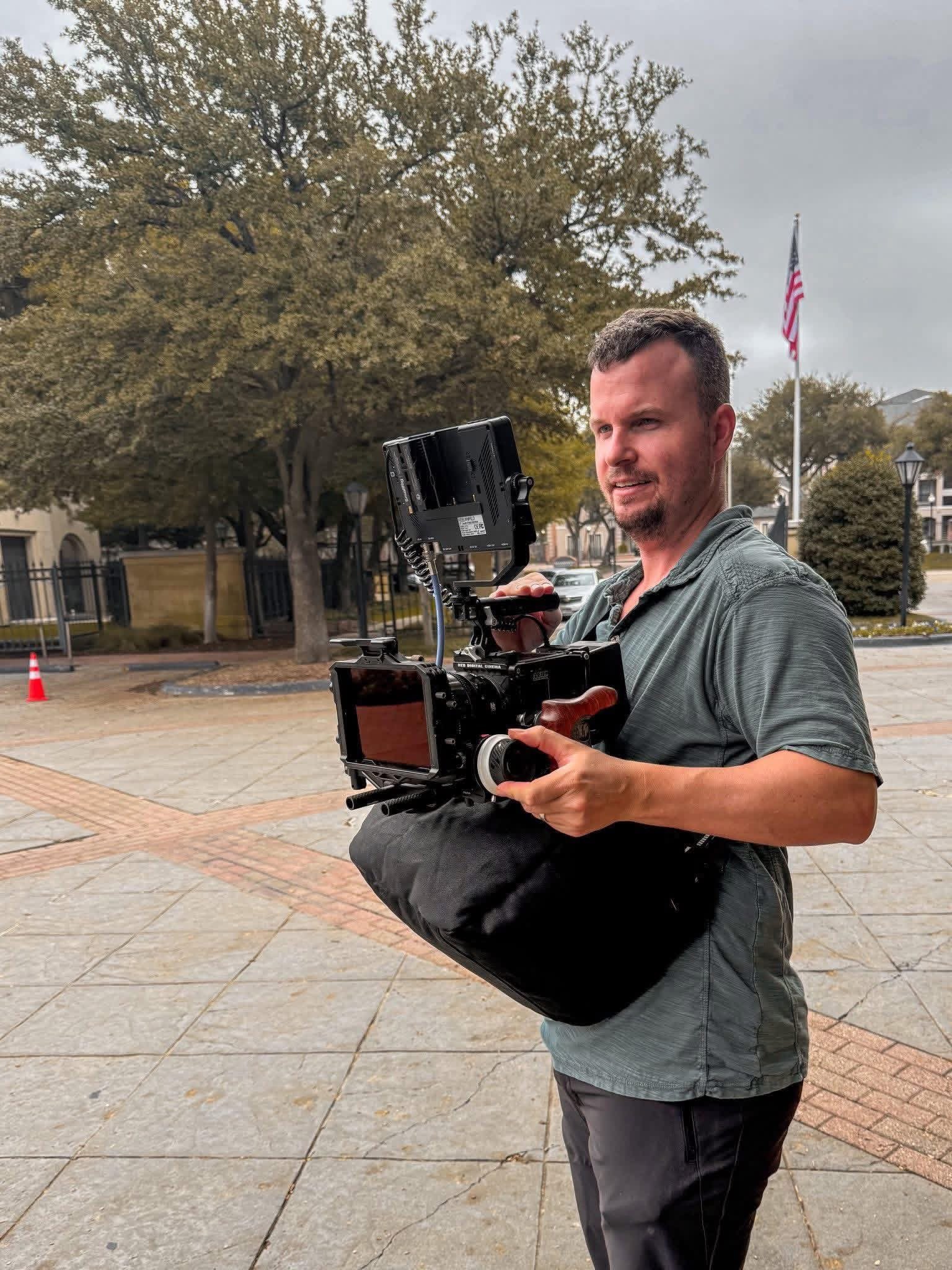 A man holding a professional video camera outdoors on a cloudy day, with trees, buildings, and an American flag in the background.