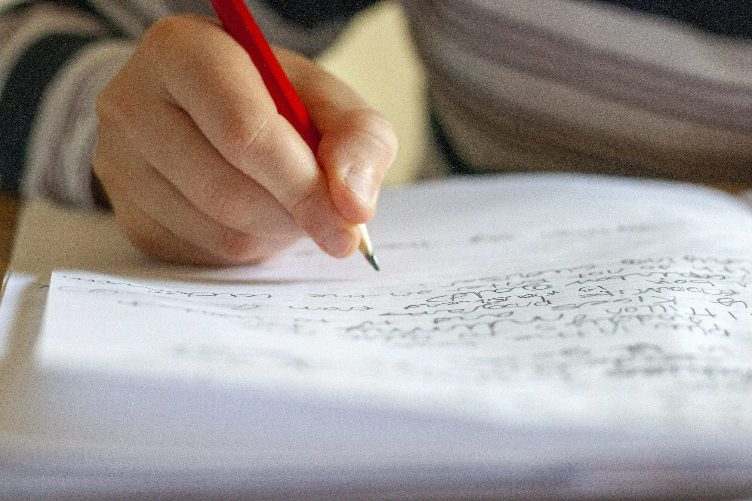 A close-up of a hand holding a red pen writing on a sheet of white paper with cursive handwriting.
