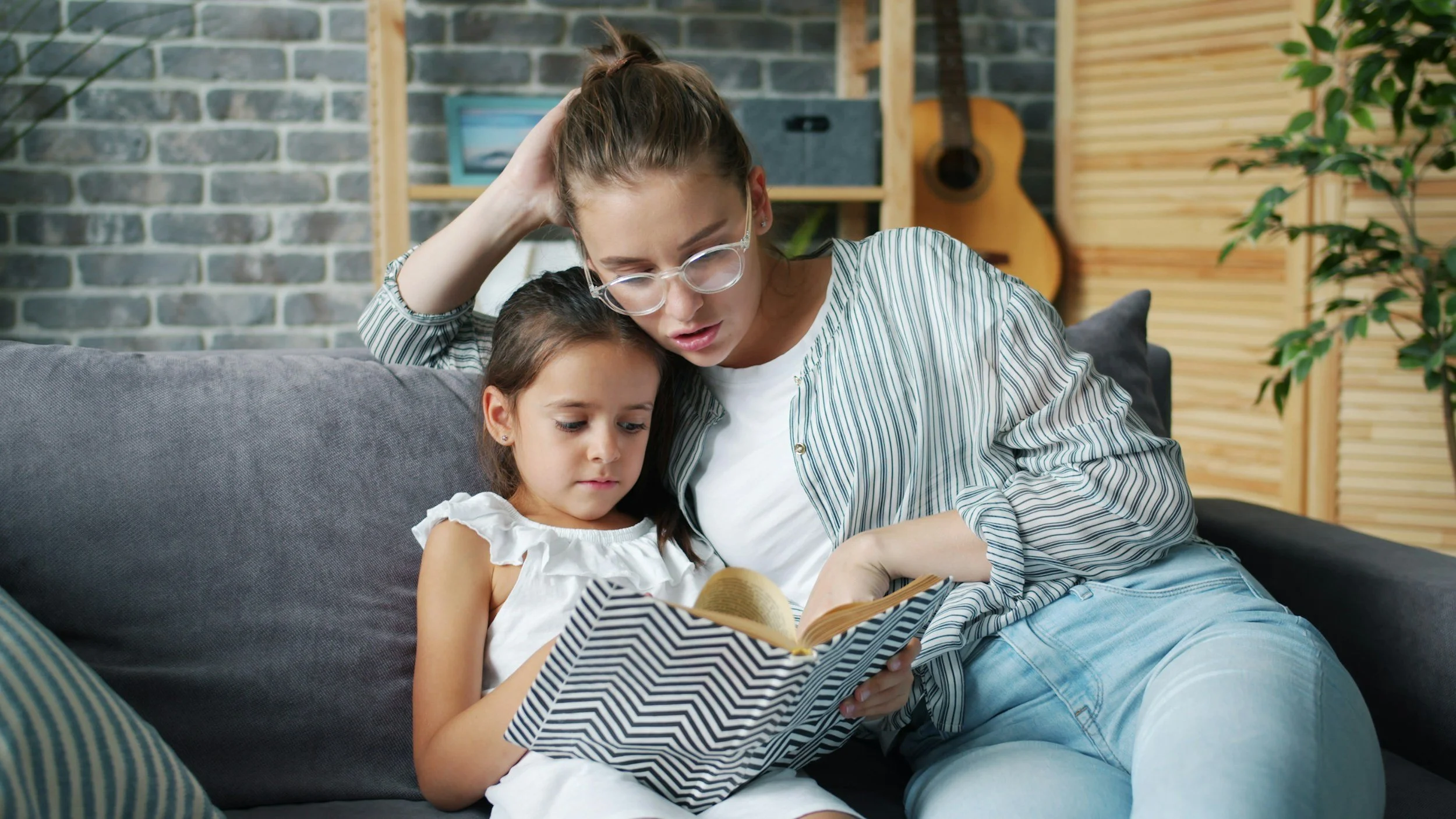 A woman and a young girl sit close together on a sofa, reading a book. The woman wears glasses and a striped shirt, while the girl has long dark hair and wears a white dress. They are in a cozy room with a brick wall, wooden shelves, a guitar, and a plant in the background.