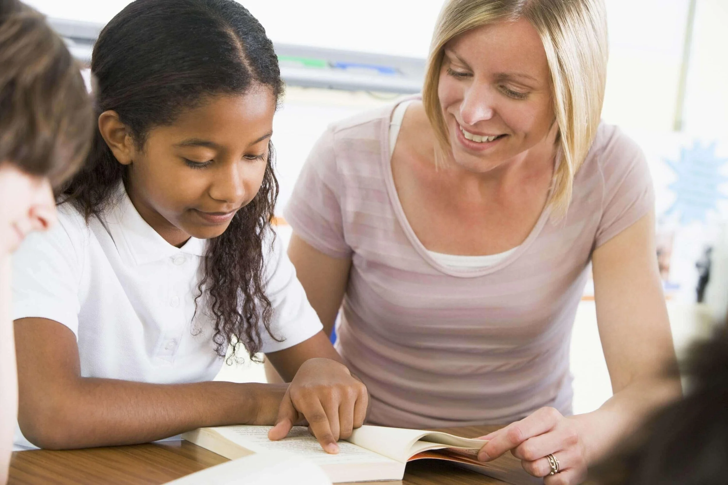 A woman with blonde hair assisting a young girl of African descent who is reading a book. They are leaning over the open book on a wooden table.