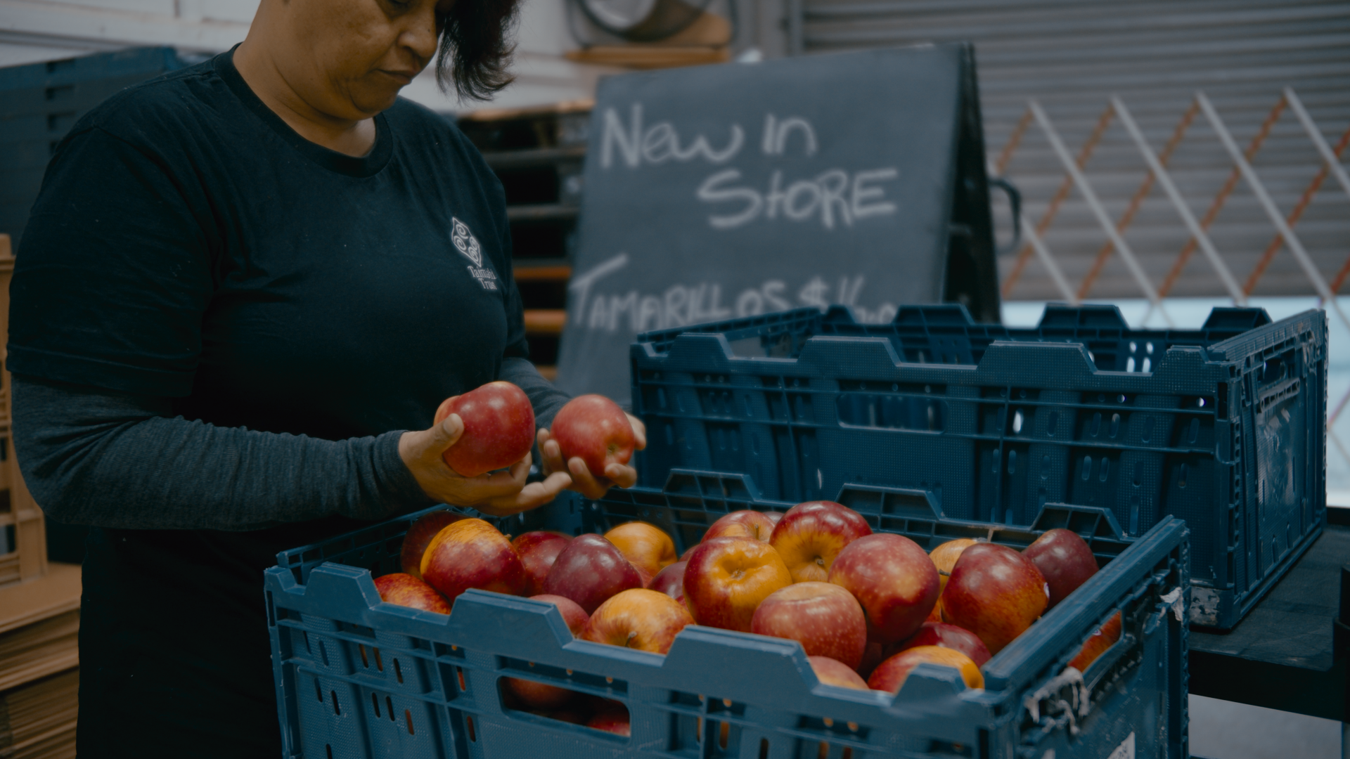 A close up of red apples in a plastic basket. A blackboard can be seen in the background, with chalk writing that says "new in store".