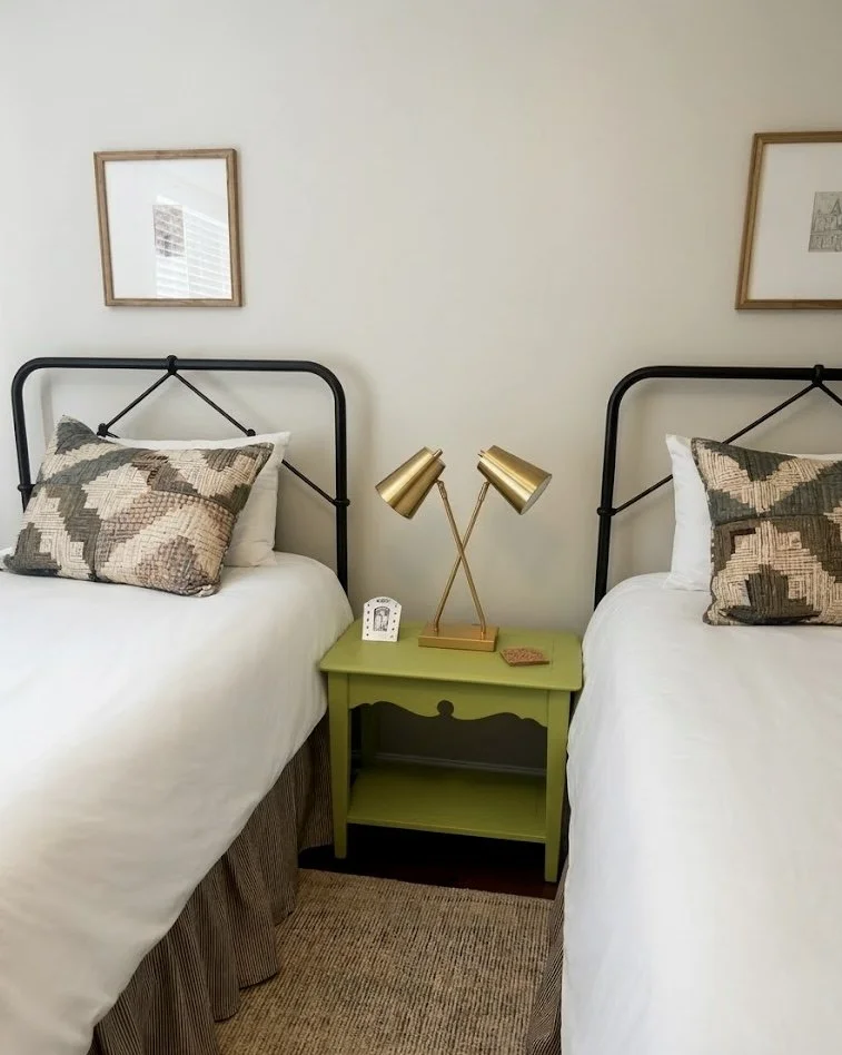 Black iron twin beds with white blankets and olive green quilted pillows, a brown striped bedskirt, brick apple green table, with gold bed lamp.