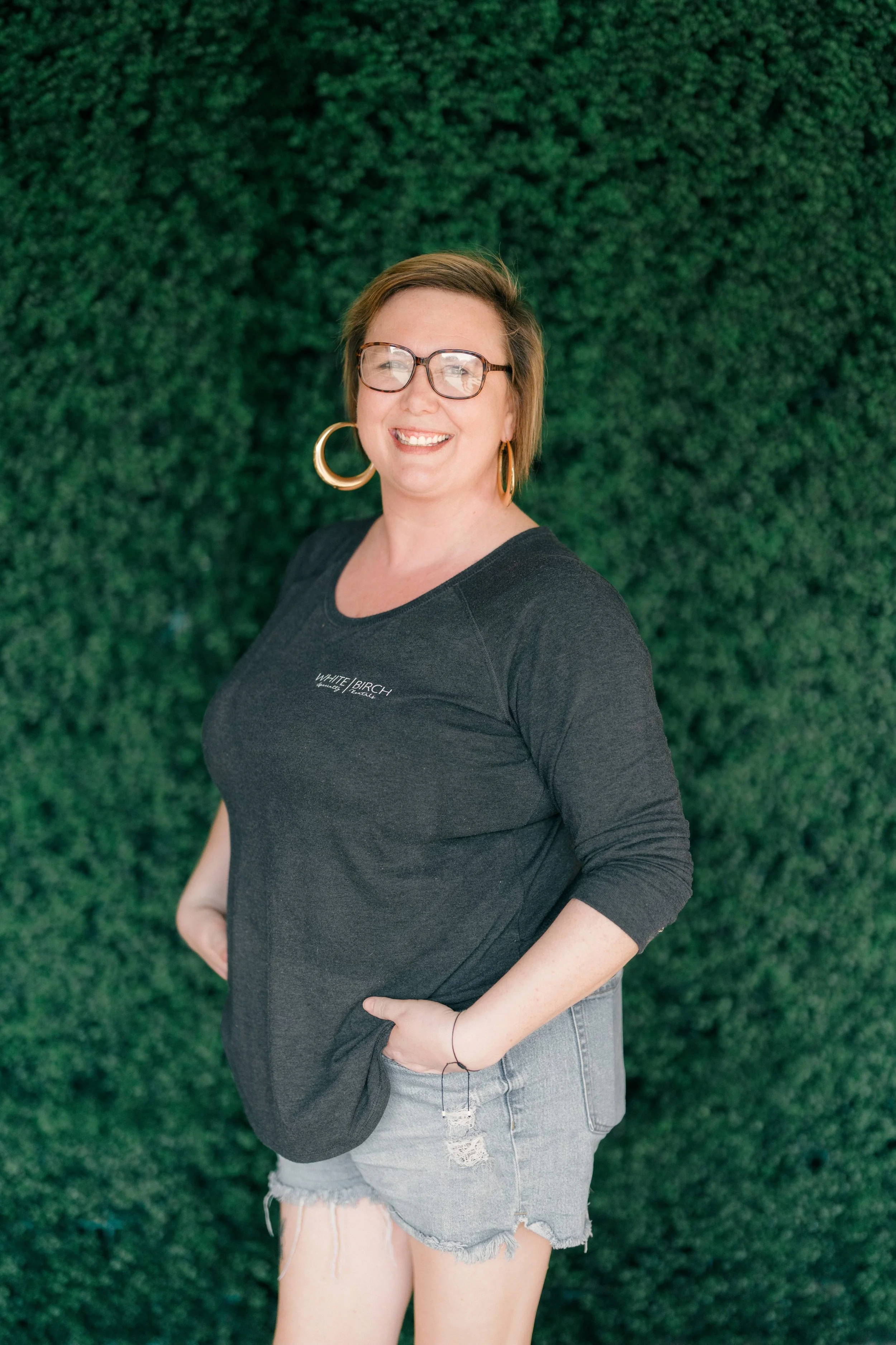 A woman standing outdoors in front of green trees, smiling, wearing glasses, a striped black and white cardigan, and a black T-shirt with white text.