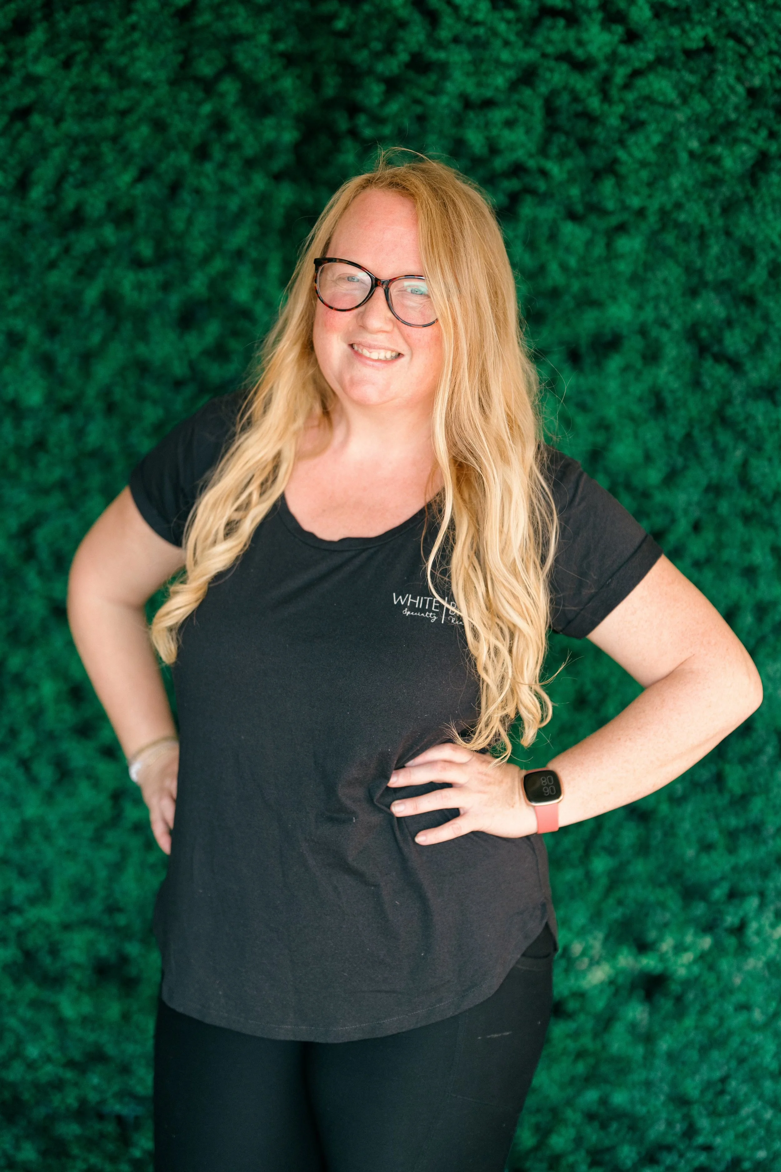 A woman with long blonde hair and glasses, wearing a black t-shirt and black pants, standing outdoors in front of green foliage and a tree with gray Spanish moss, smiling at the camera.